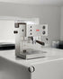 Lelit Kate PL82T espresso machine with built-in grinder on white countertop, featuring stainless steel body, control panel, and transparent bean hopper
