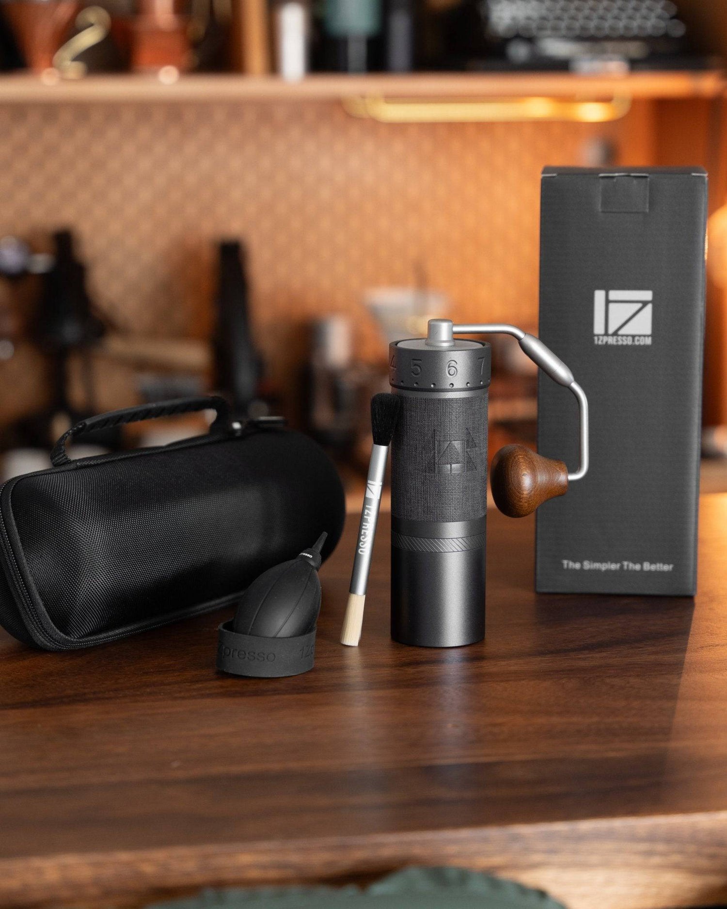 1zpresso J MAXS manual coffee grinder with accessories including carry case, cleaning brush, and product box displayed on wooden counter with coffee equipment in background