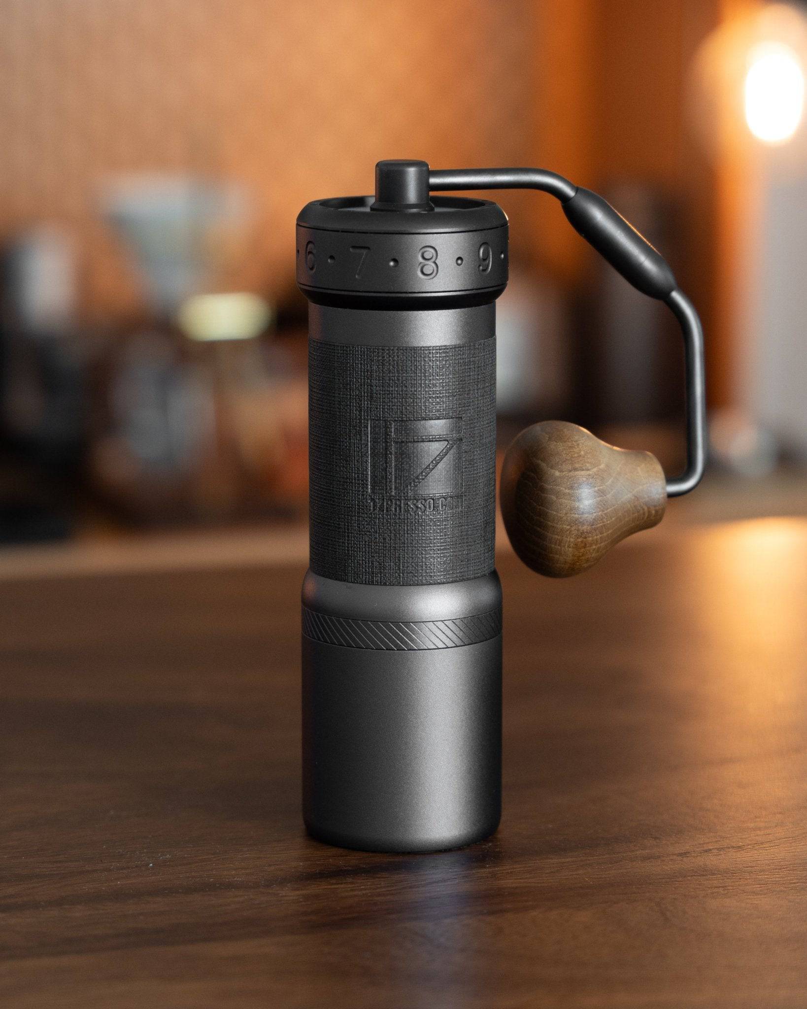 1Zpresso K-Ultra manual coffee grinder with foldable handle, sleek black design, and wooden knob on wooden table with blurred coffee equipment in background