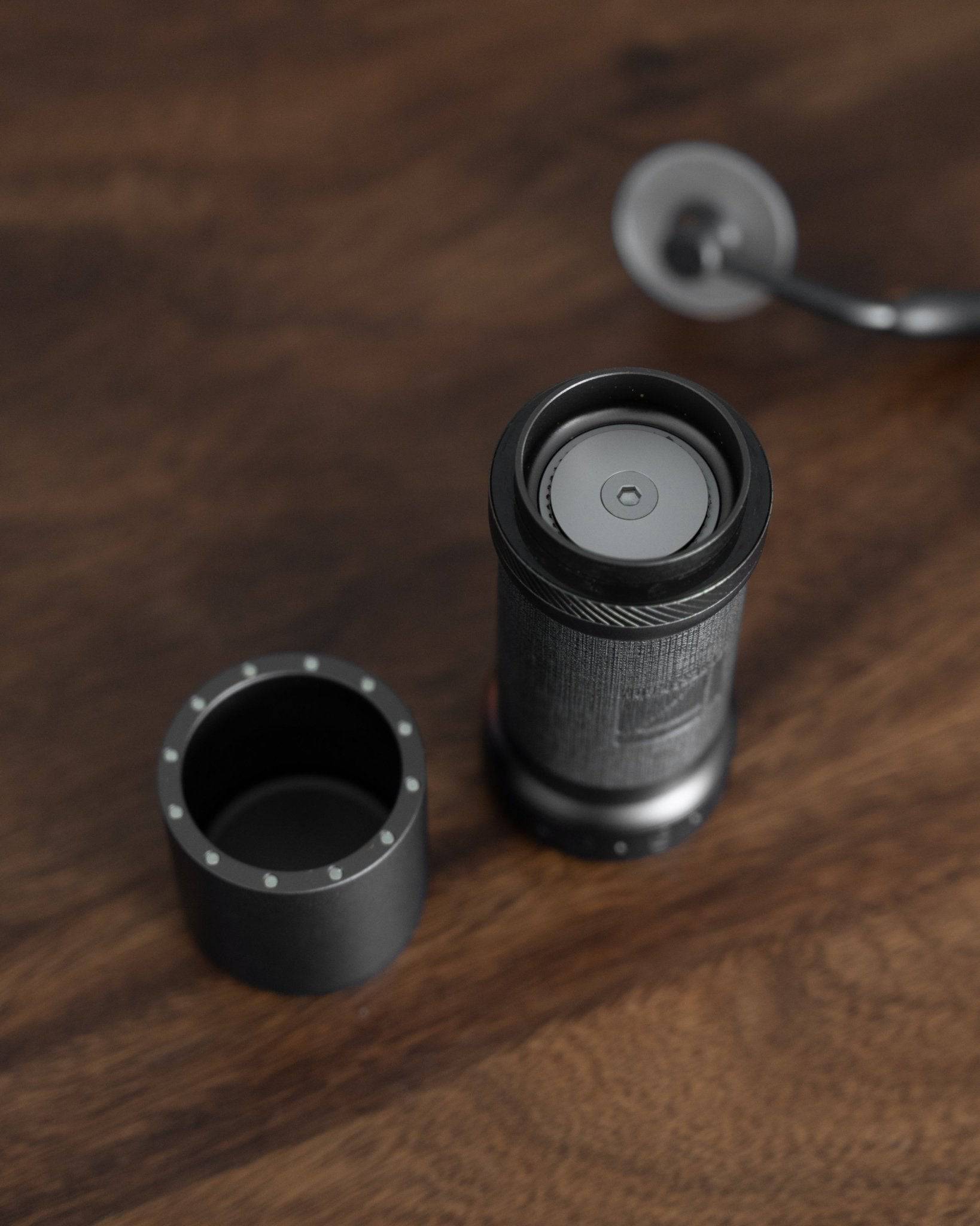 1Zpresso K-Ultra manual coffee grinder disassembled on wooden surface, showing sleek black cylindrical body, adjustable grind setting, and detachable components for easy cleaning and portability