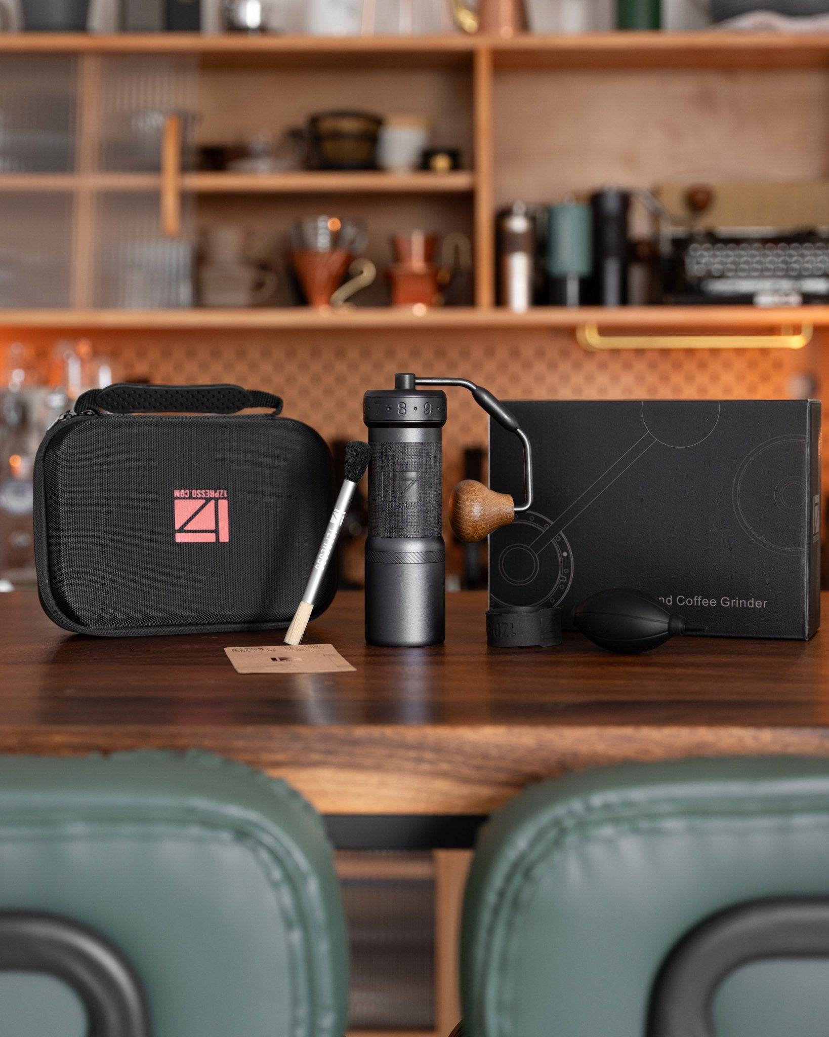1Zpresso K-Ultra hand coffee grinder set on wooden table in kitchen, including black grinder, storage case, and accessories, with shelves of coffee equipment in background