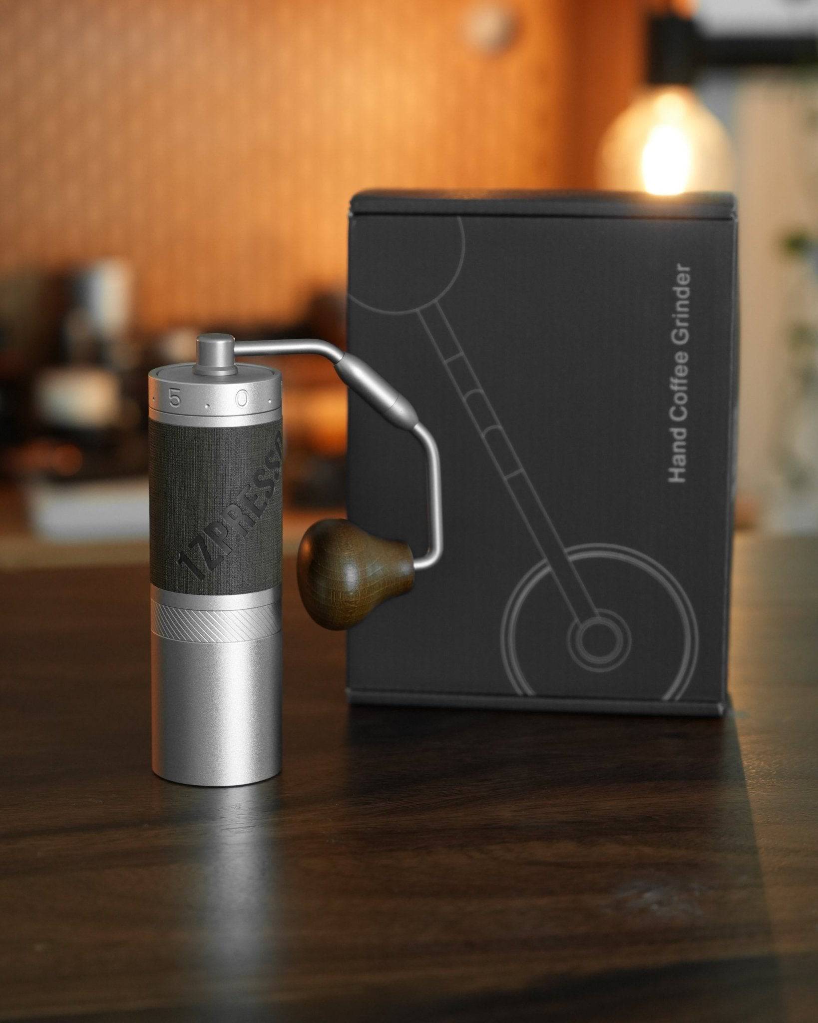 1Zpresso X-Pro-S manual coffee grinder with sleek aluminum body and precise adjustment settings, displayed next to its stylish black packaging featuring minimalist grinder illustration on wooden surface