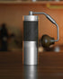 1Zpresso X-Pro-S manual coffee grinder with sleek aluminum body, black textured grip, and ergonomic wooden handle on wooden surface, showcasing its portable and professional design for coffee enthusiasts