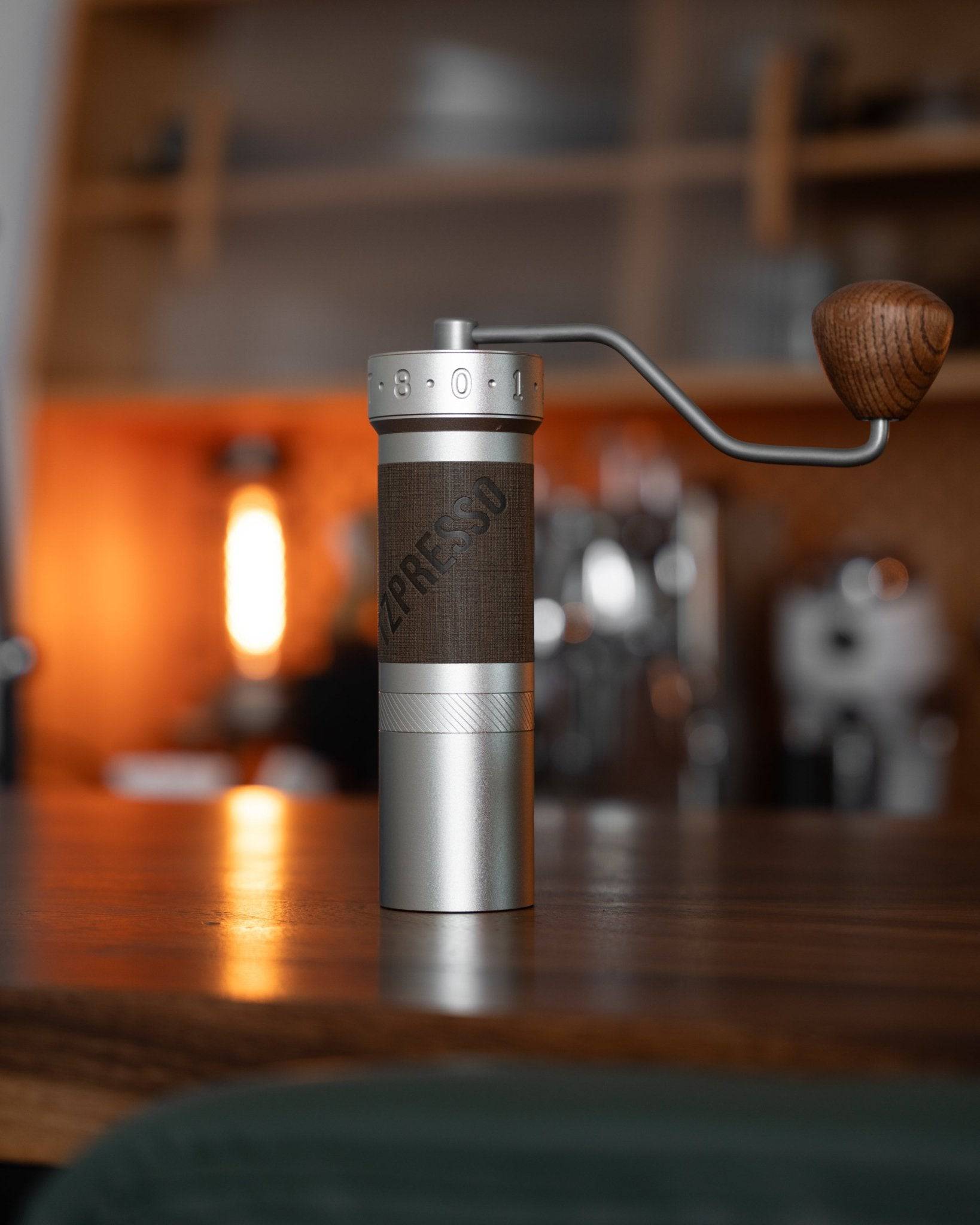 1zpresso ZP6 manual coffee grinder on wooden counter, featuring silver and gray body with wooden handle. Warm lighting in background showcases premium design for pour-over coffee enthusiasts.