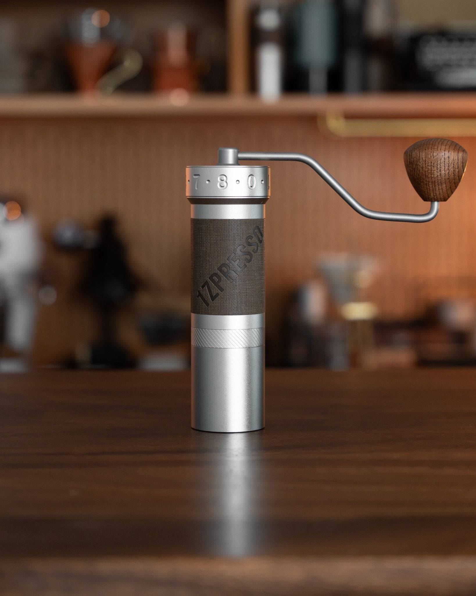 1zpresso ZP6 manual coffee grinder on wooden table, featuring silver body, wood handle, and adjustable grind settings, with blurred kitchen background