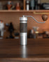 1zpresso ZP6 manual coffee grinder on wooden table, featuring silver body, wood handle, and adjustable grind settings, with blurred kitchen background
