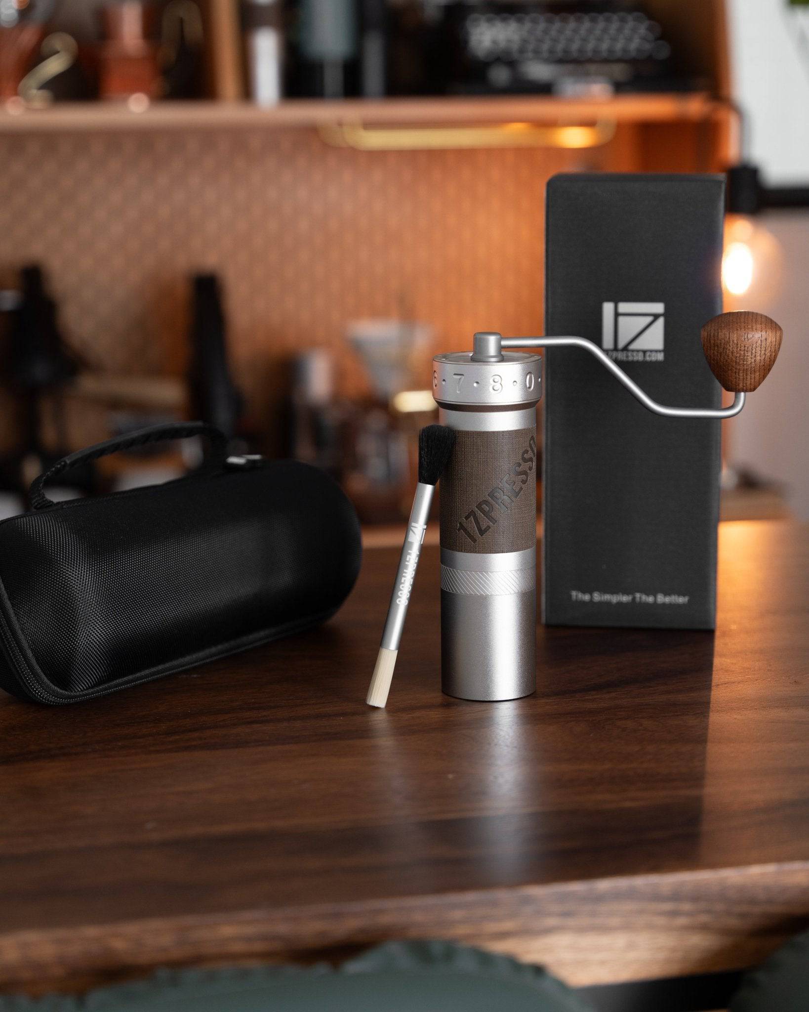 1zpresso ZP6 manual coffee grinder with wooden handle, displayed on wooden table alongside its packaging, cleaning brush, and carrying case in a cozy coffee shop setting