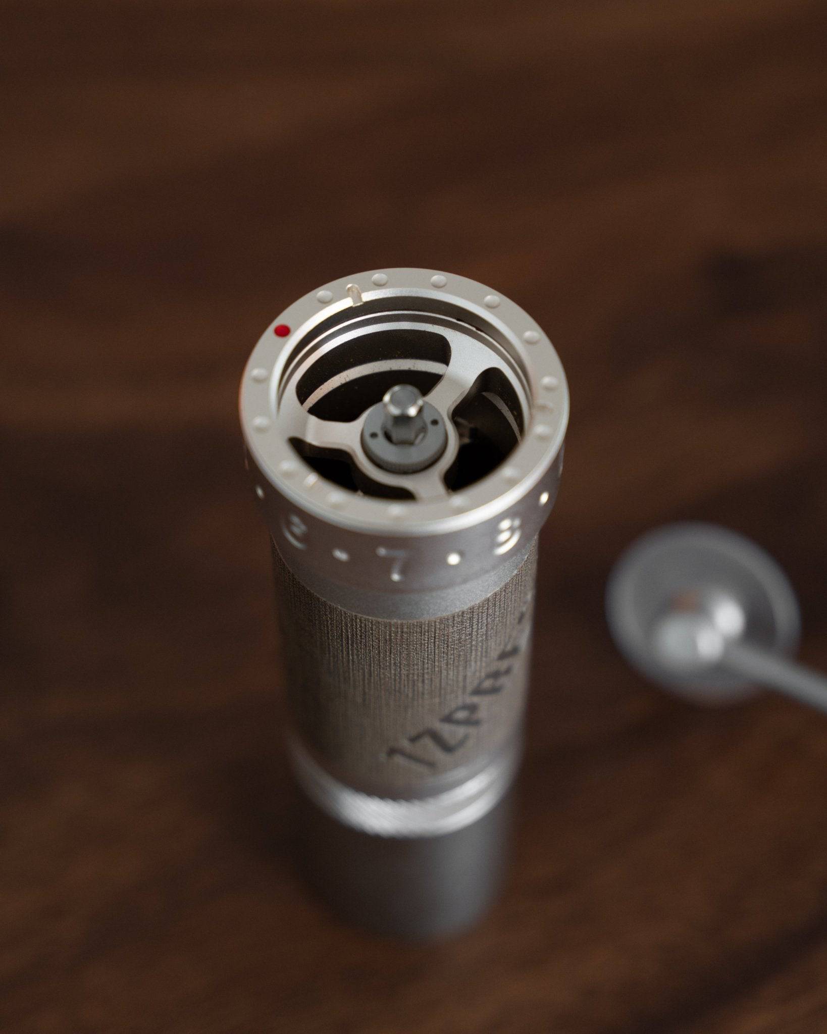Close-up view of 1zpresso ZP6 manual coffee grinder, showing the top grinding mechanism and silver cylindrical body, with a wooden background and part of the grinder handle visible.