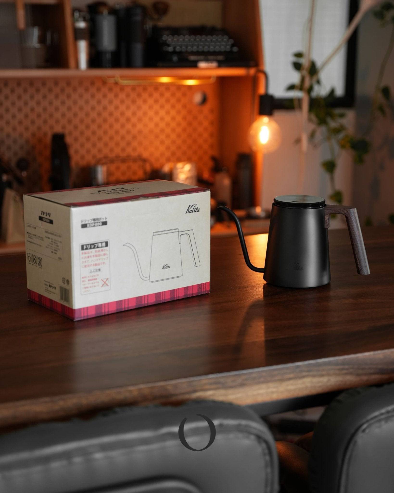 Kalita KDP-800 black walnut wood pattern coffee pour-over kettle with product box on wooden counter in warm, cozy kitchen setting with ambient lighting