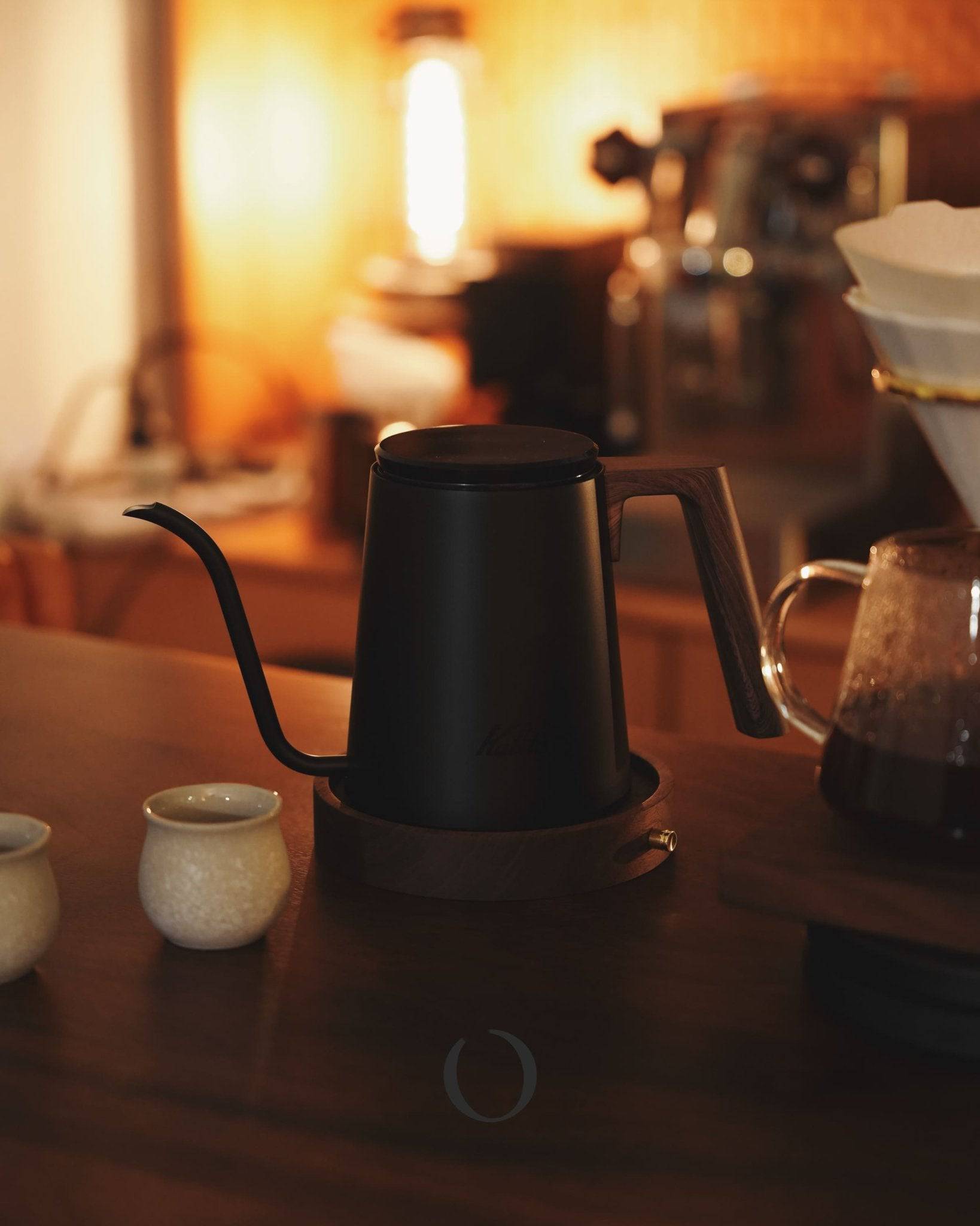 Elegant black Kalita KDP-800 pour-over kettle on wooden table with warm lighting, surrounded by coffee brewing accessories, showcasing its sleek design and practical functionality for coffee enthusiasts