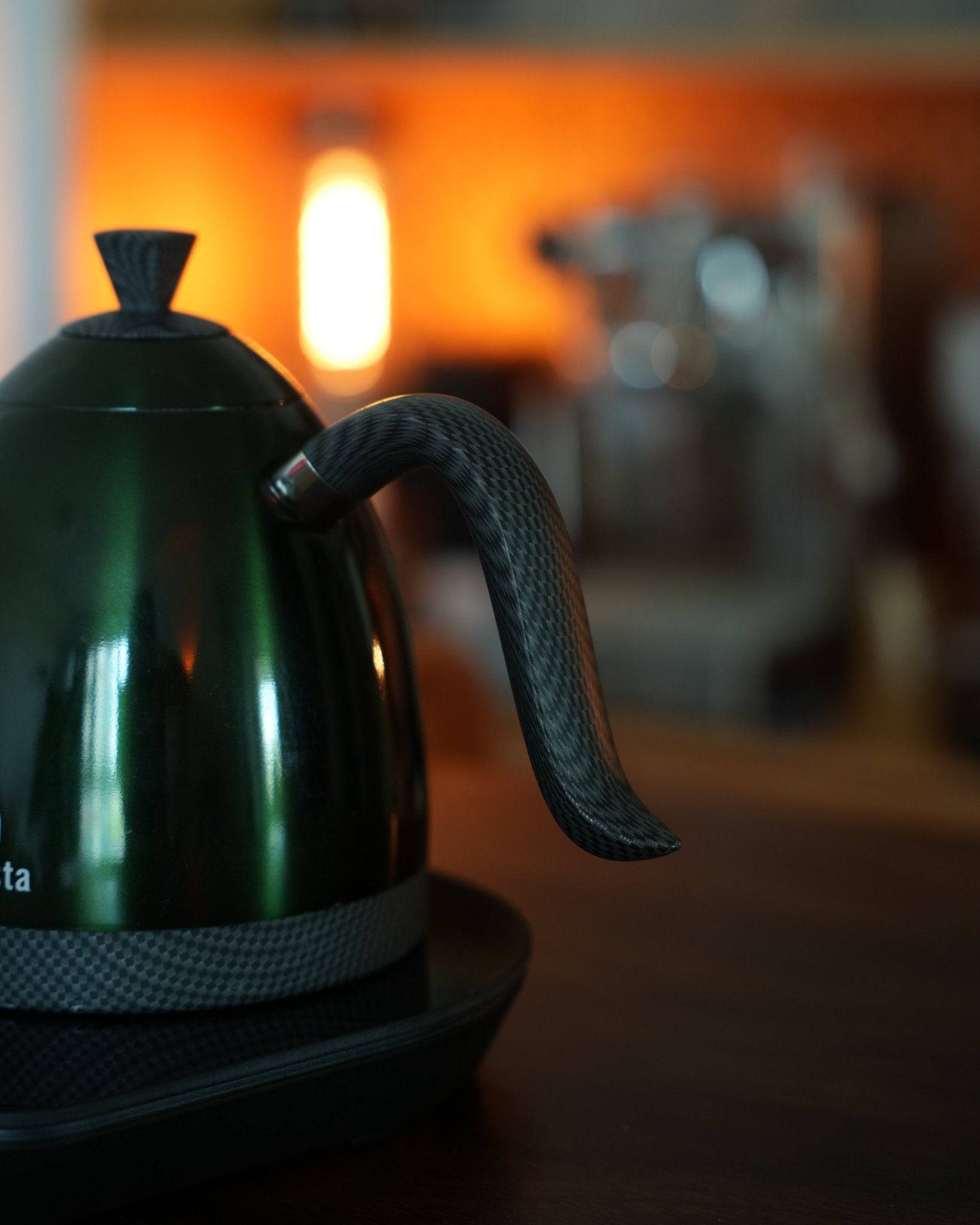 Brewista Artisan temperature-controlled kettle with gooseneck spout in dark color on wooden surface, warm ambient lighting, blurred coffee equipment background
