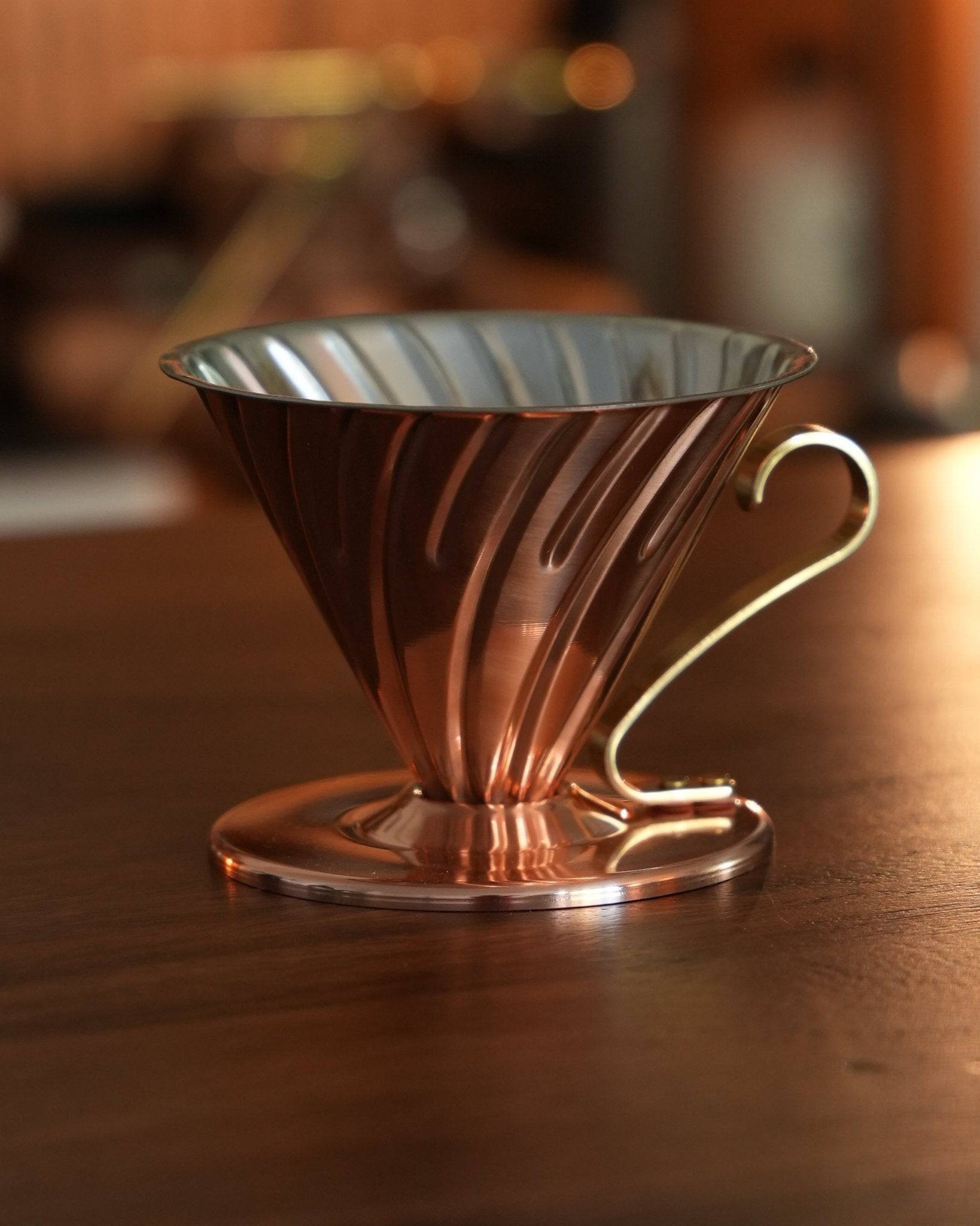 Copper HARIO V60 02 pour-over coffee dripper with conical design and spiral ridges, featuring a brass handle, on wooden surface in warm lighting