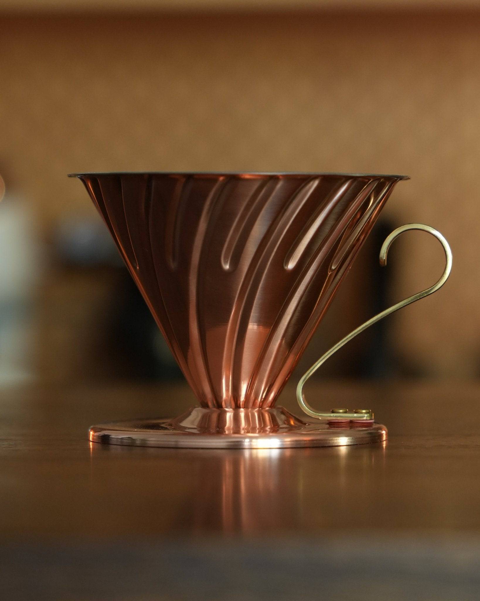 HARIO V60 02 copper coffee dripper with conical design and brass handle, showcasing elegant Japanese craftsmanship for premium pour-over brewing, sitting on a wooden surface in warm lighting