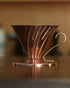 HARIO V60 02 copper coffee dripper with conical design and brass handle, showcasing elegant Japanese craftsmanship for premium pour-over brewing, sitting on a wooden surface in warm lighting