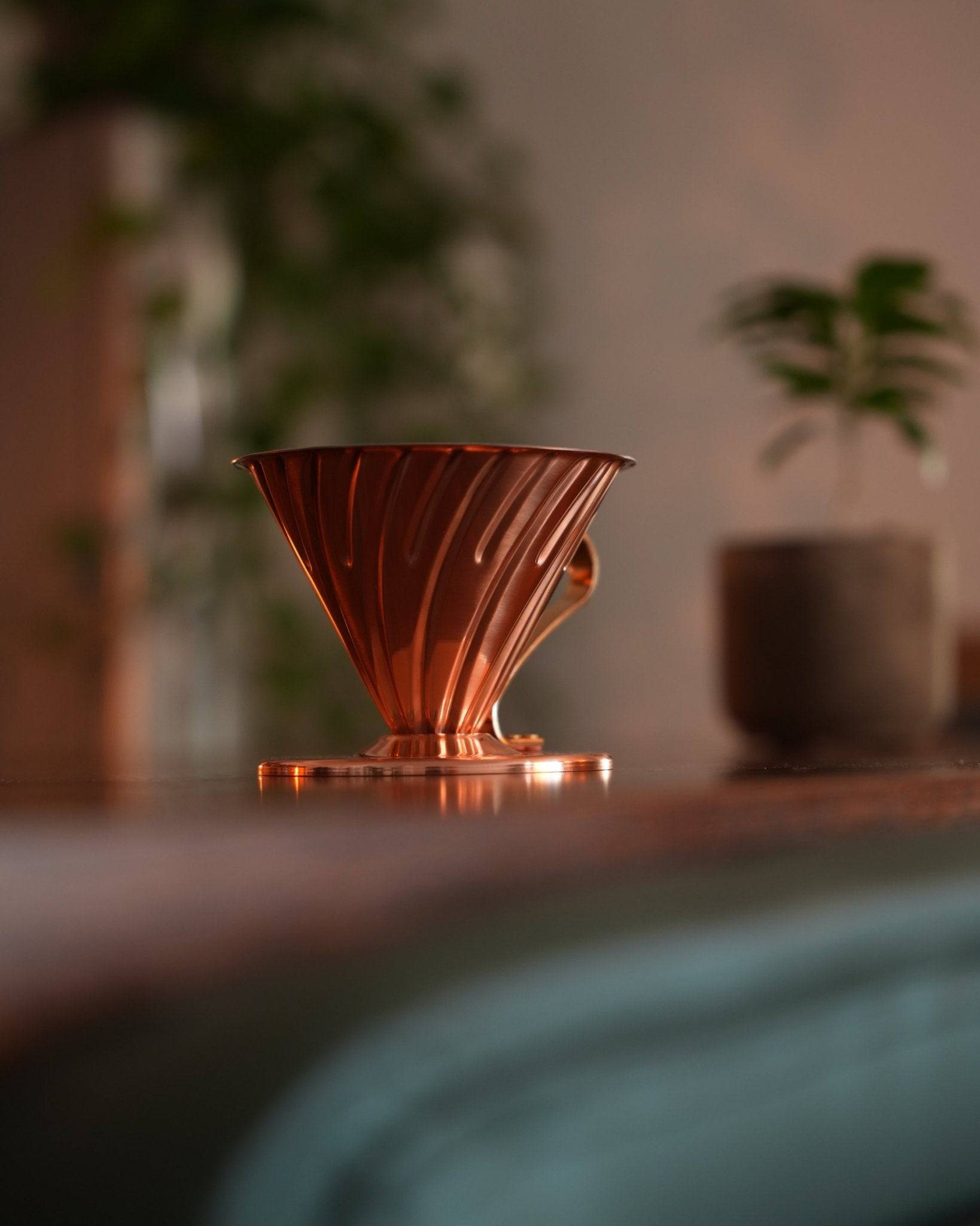 HARIO V60 02 copper pour-over coffee dripper on wooden surface, surrounded by blurred plants, showcasing its elegant design and warm copper hue in a cozy home setting