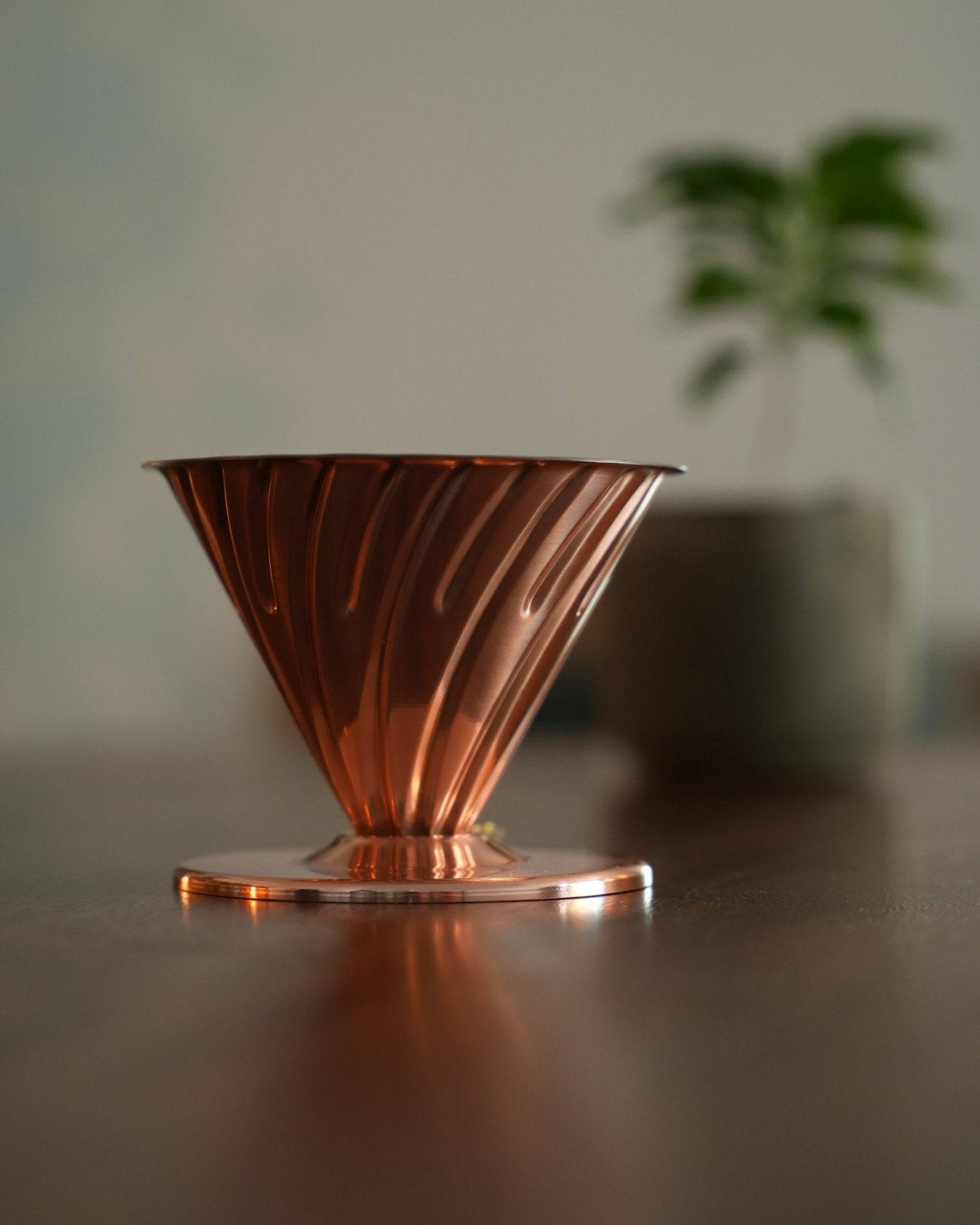 HARIO V60 02 copper pour-over coffee dripper on wooden surface, conical shape with spiral ridges, reflecting warm light, green plant blurred in background