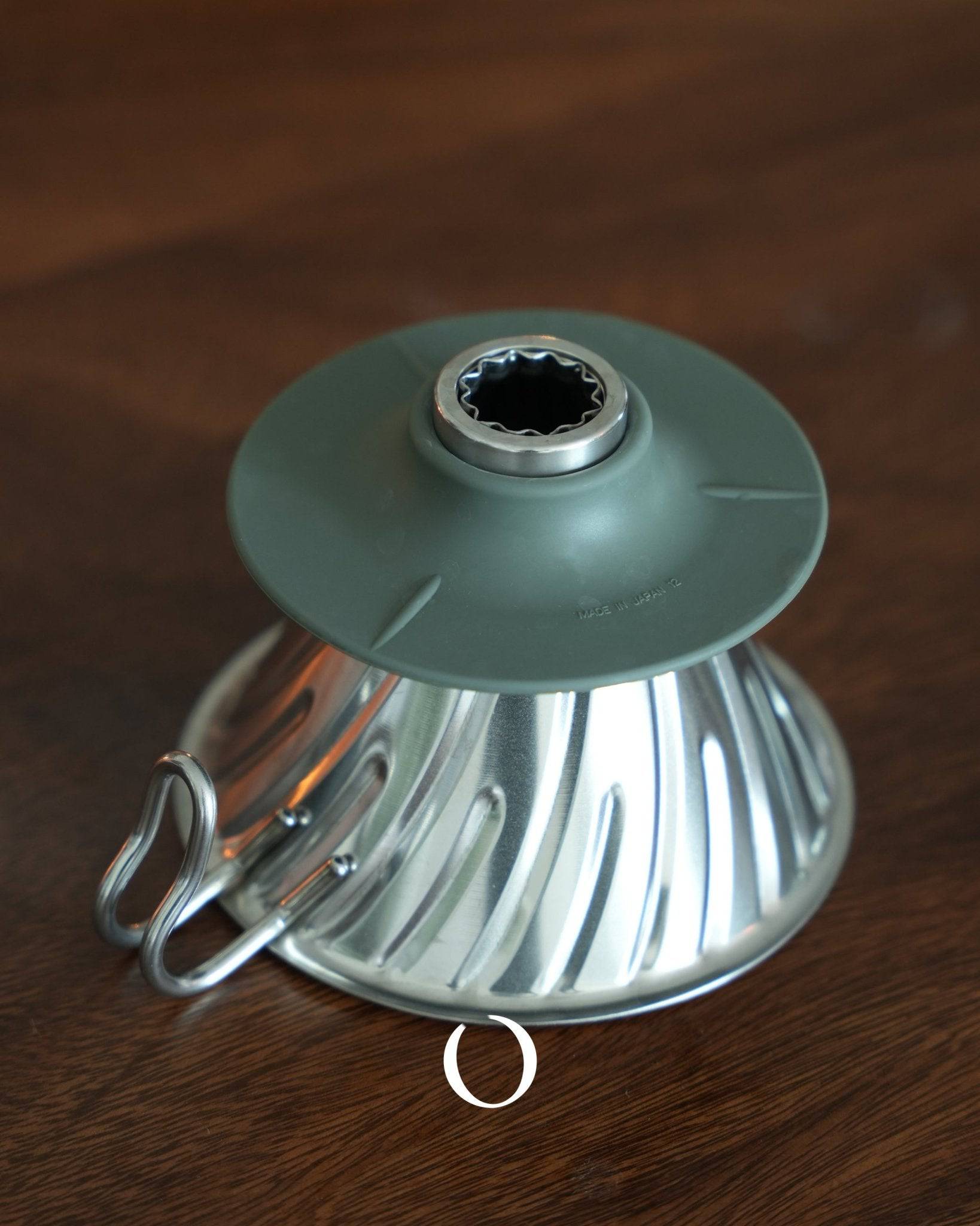 HARIO V60 02 stainless steel coffee dripper in silver, featuring a conical design with spiral ribs and a silicone handle, placed on a wooden surface, showcasing its durable and easy-to-clean construction for optimal coffee extraction.