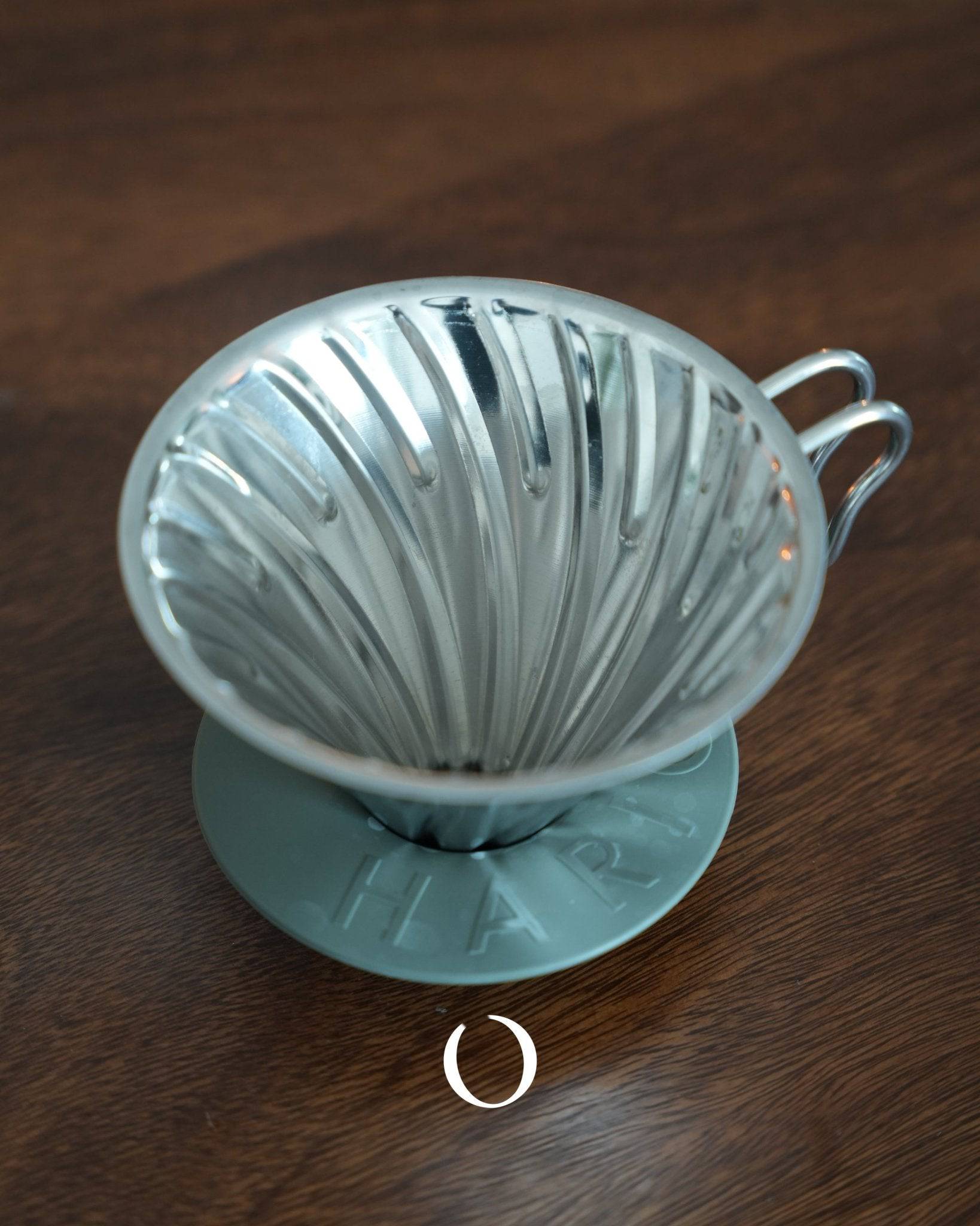 HARIO V60 02 stainless steel pour-over coffee dripper in silver, featuring classic conical design with spiral ribs, placed on wooden surface, showcasing its durable and easy-to-clean construction for optimal coffee extraction