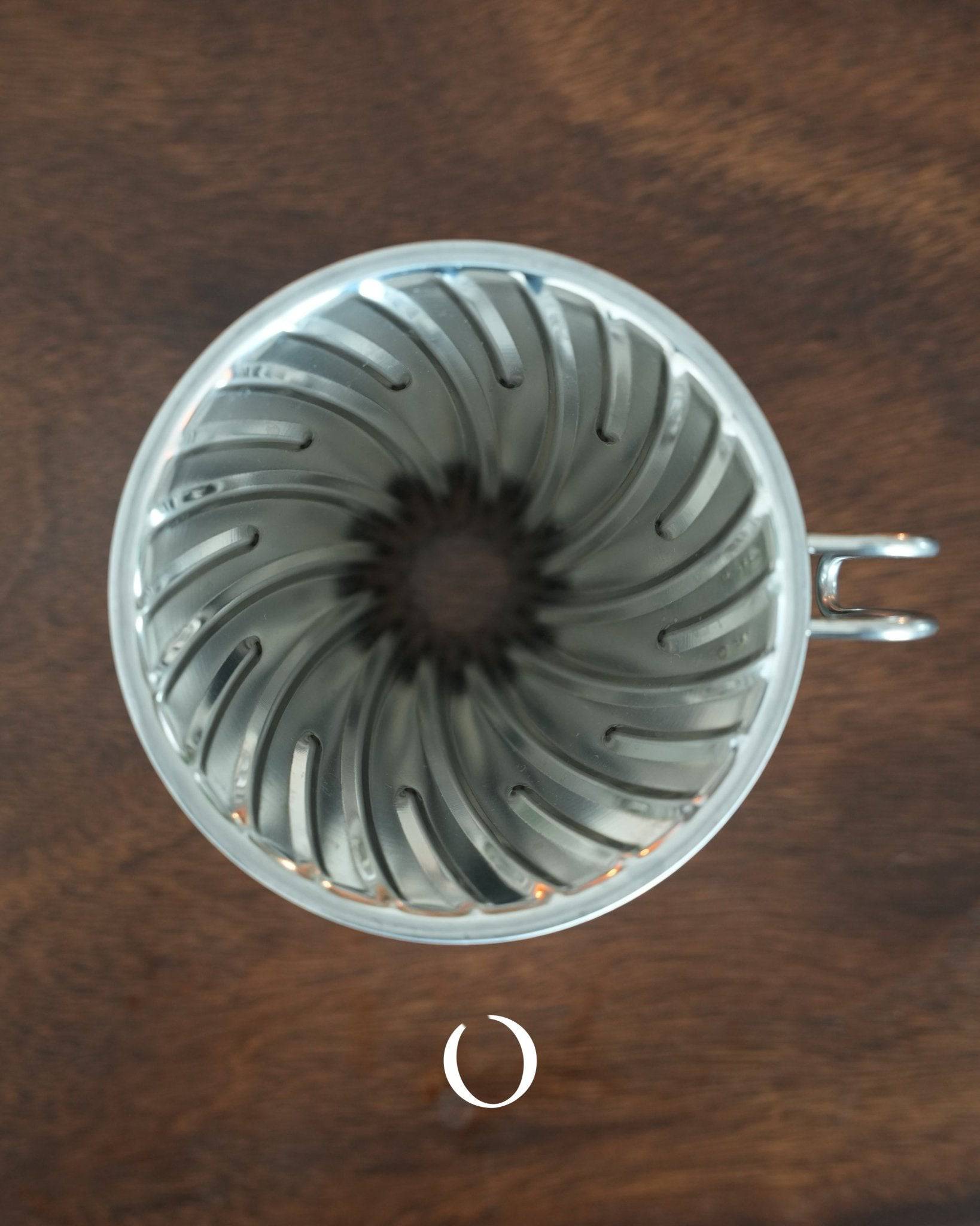 HARIO V60 02 stainless steel pour-over coffee dripper in silver, featuring a classic conical design with spiral ribs, viewed from above on a wooden surface, showcasing its durable and easy-to-clean construction for optimal coffee extraction.