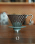 HARIO V60 02 stainless steel coffee dripper in silver, featuring a conical shape with spiral ribs, placed on a wooden surface with a blurred kitchen background, showcasing its durable and easy-to-clean design for optimal coffee brewing