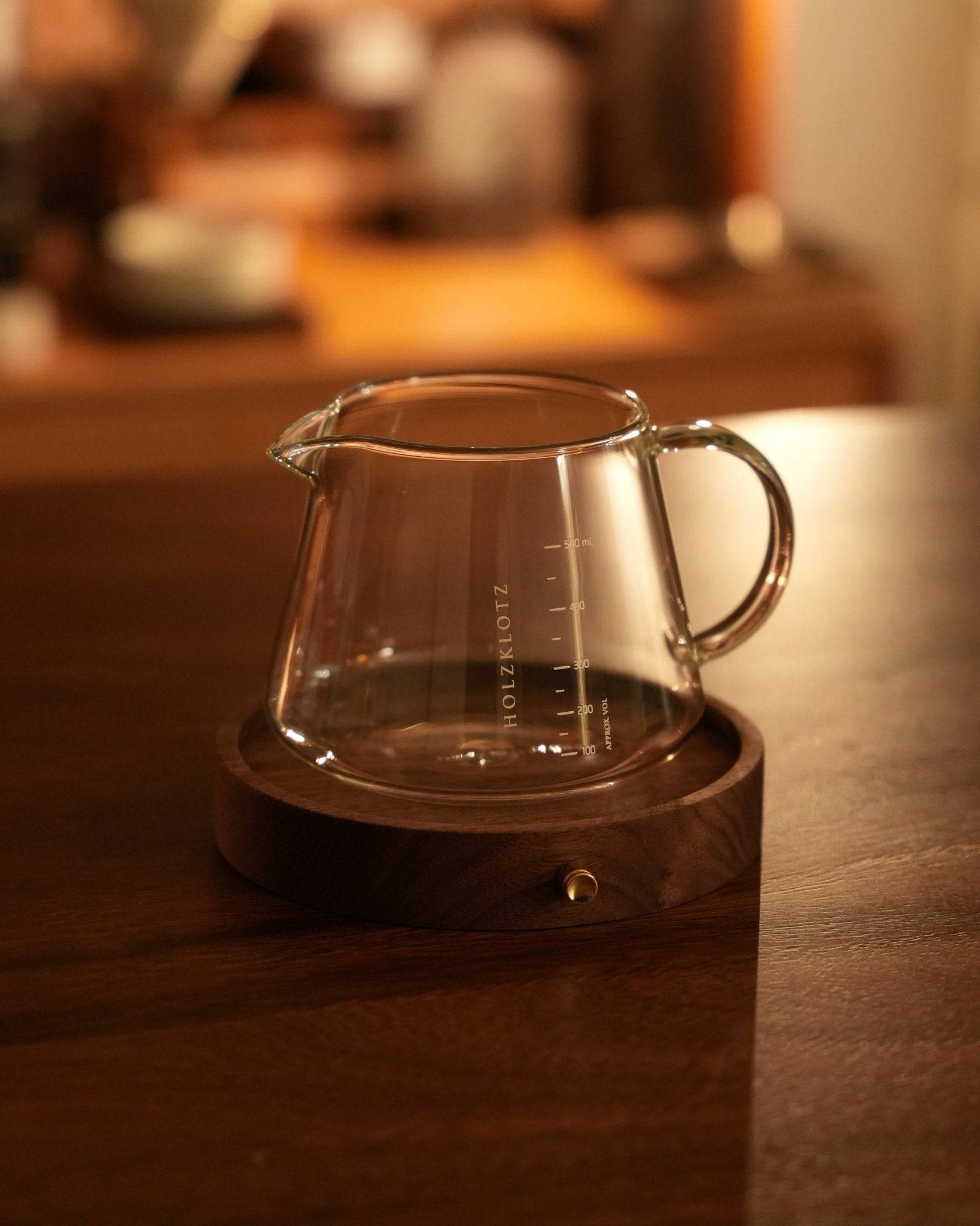 Glass coffee server on dark wooden Holkzlotz stand, warm ambient lighting, rustic coffee bar setting, premium barista equipment, artisanal coffee brewing accessory