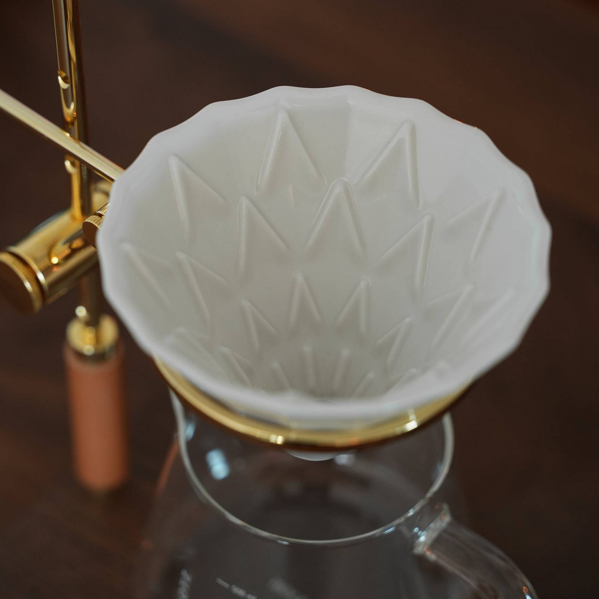 Elegant pour-over coffee set with white ceramic filter, gold-plated stand, and glass carafe on dark wood surface, showcasing Holzklotz&