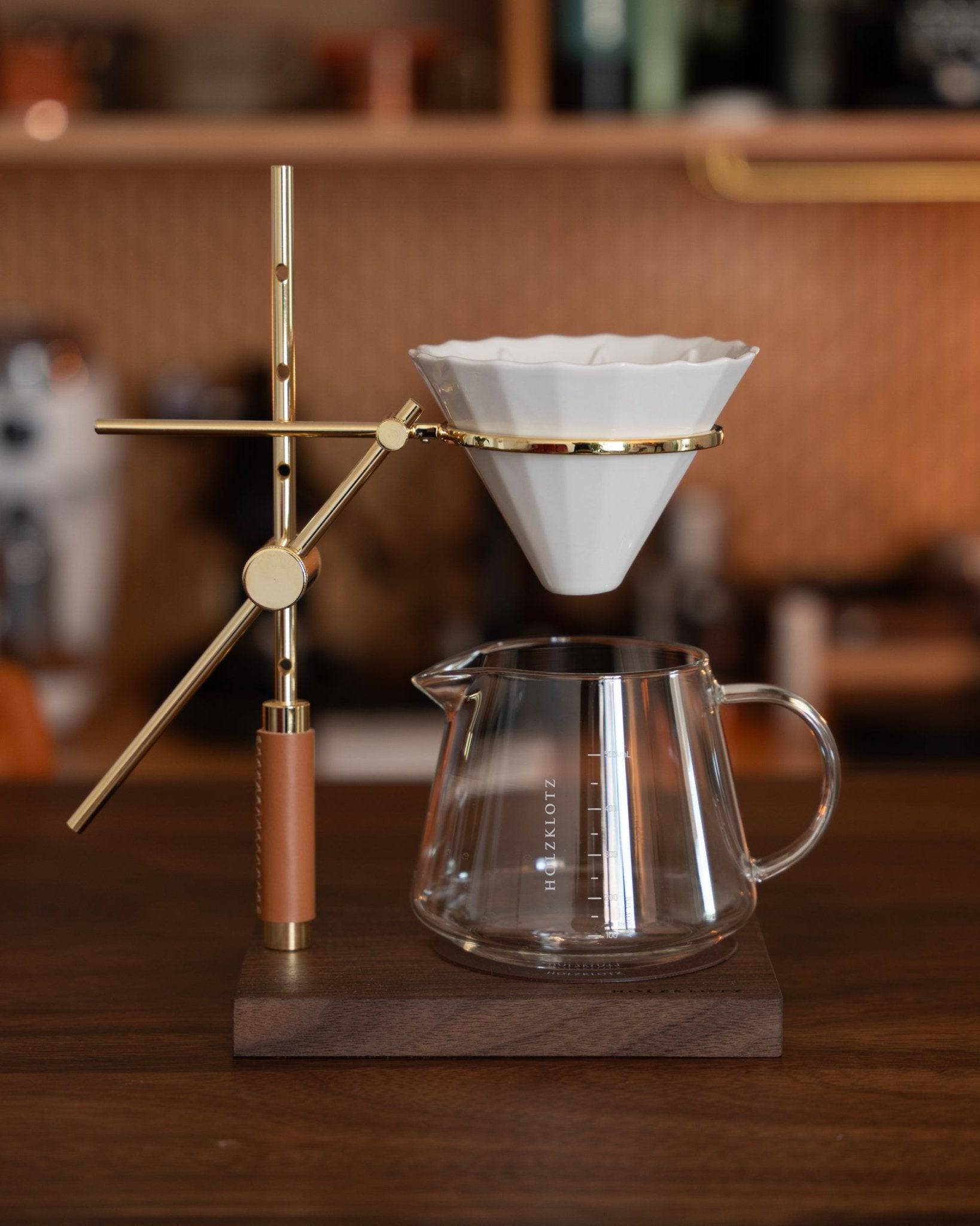 Elegant pour-over coffee set featuring gold-plated stand, white ceramic dripper, and glass carafe on wooden base, showcasing Holzklotz&