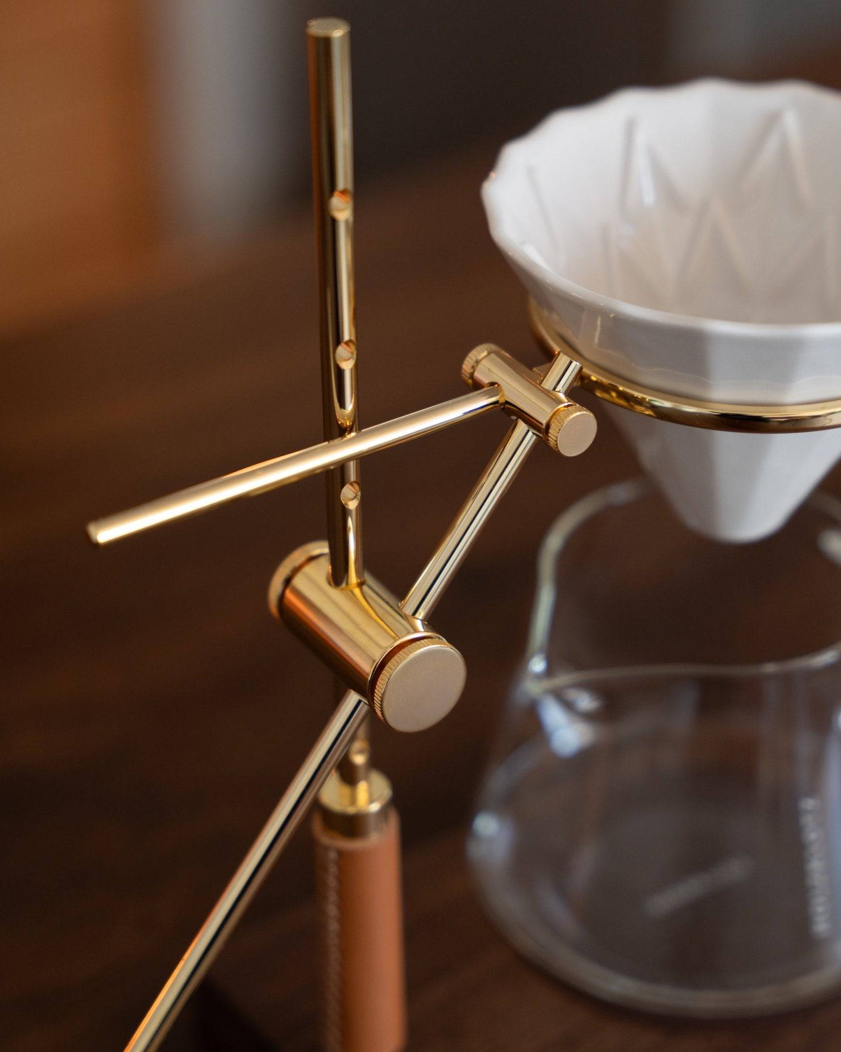 Elegant gold-plated pour-over coffee stand with adjustable arms, holding a white ceramic dripper over a glass carafe, showcasing Holzklotz&