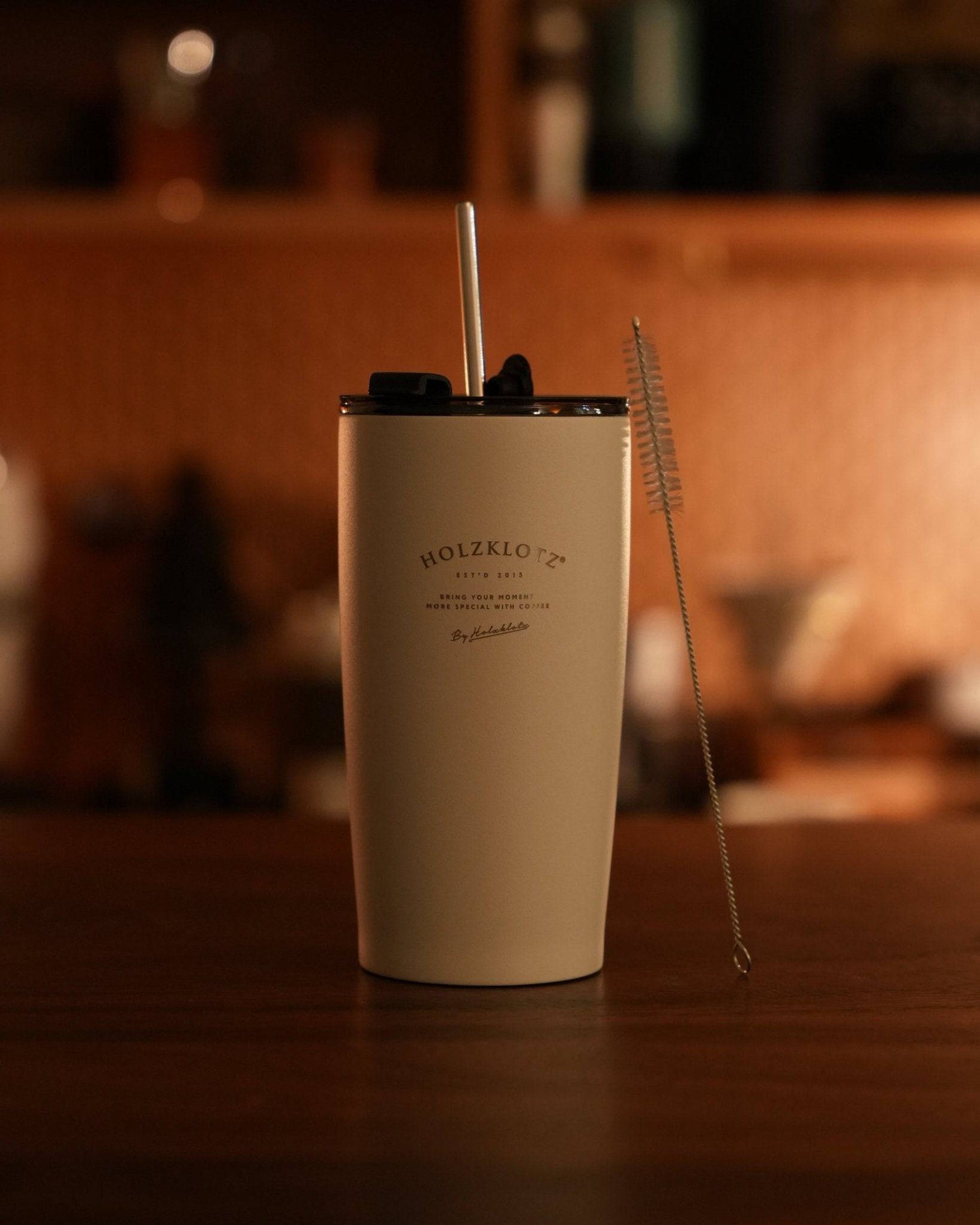 Holzklotz vacuum insulated tumbler 500ml in beige color with straw and cleaning brush on wooden table, warm ambient lighting, cozy home atmosphere