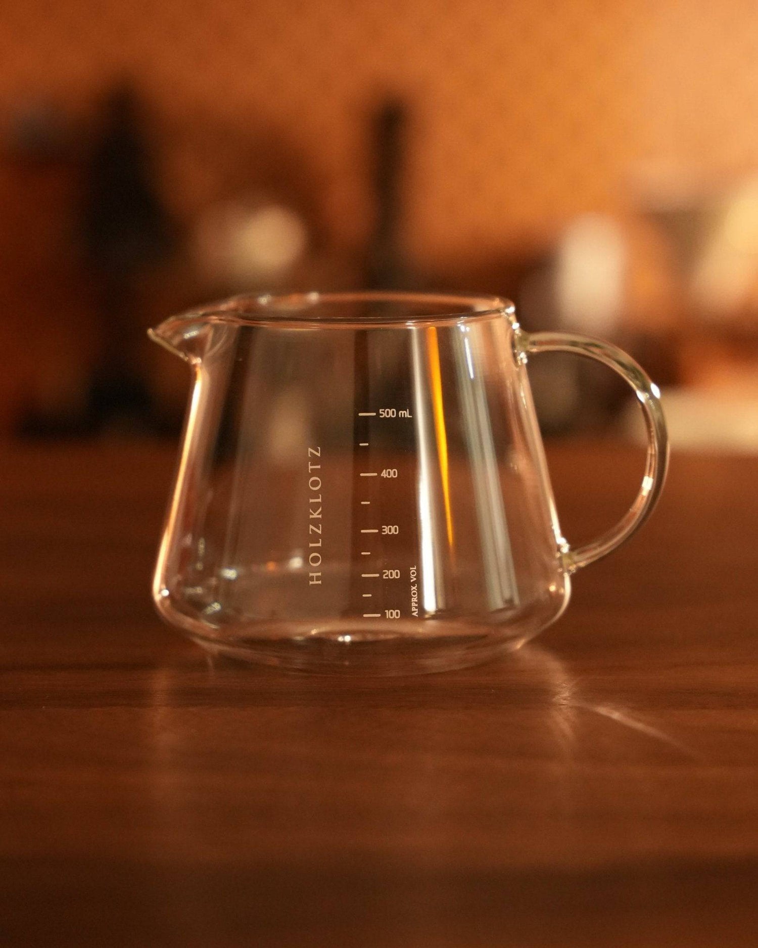 Glass coffee server with Holzklotz branding and 500ml capacity, featuring measurement markings and a pour spout, placed on a wooden surface in a warm, ambient setting