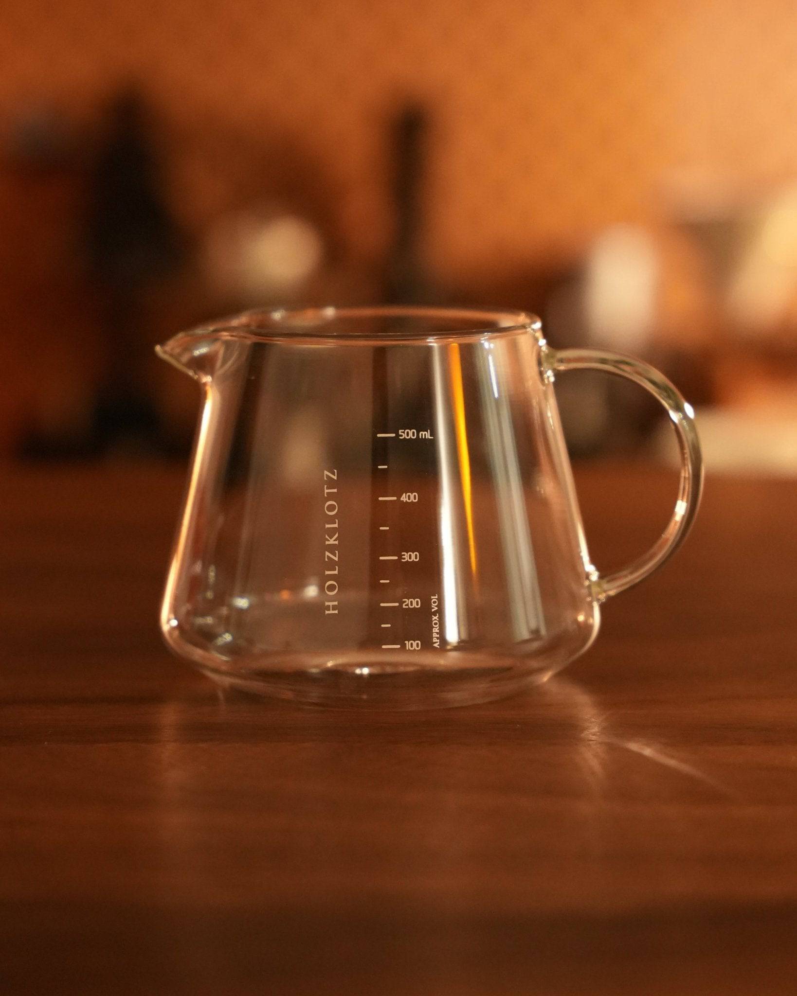 Glass coffee server with Holzklotz branding and 500ml capacity, featuring measurement markings and a pour spout, placed on a wooden surface in a warm, ambient setting