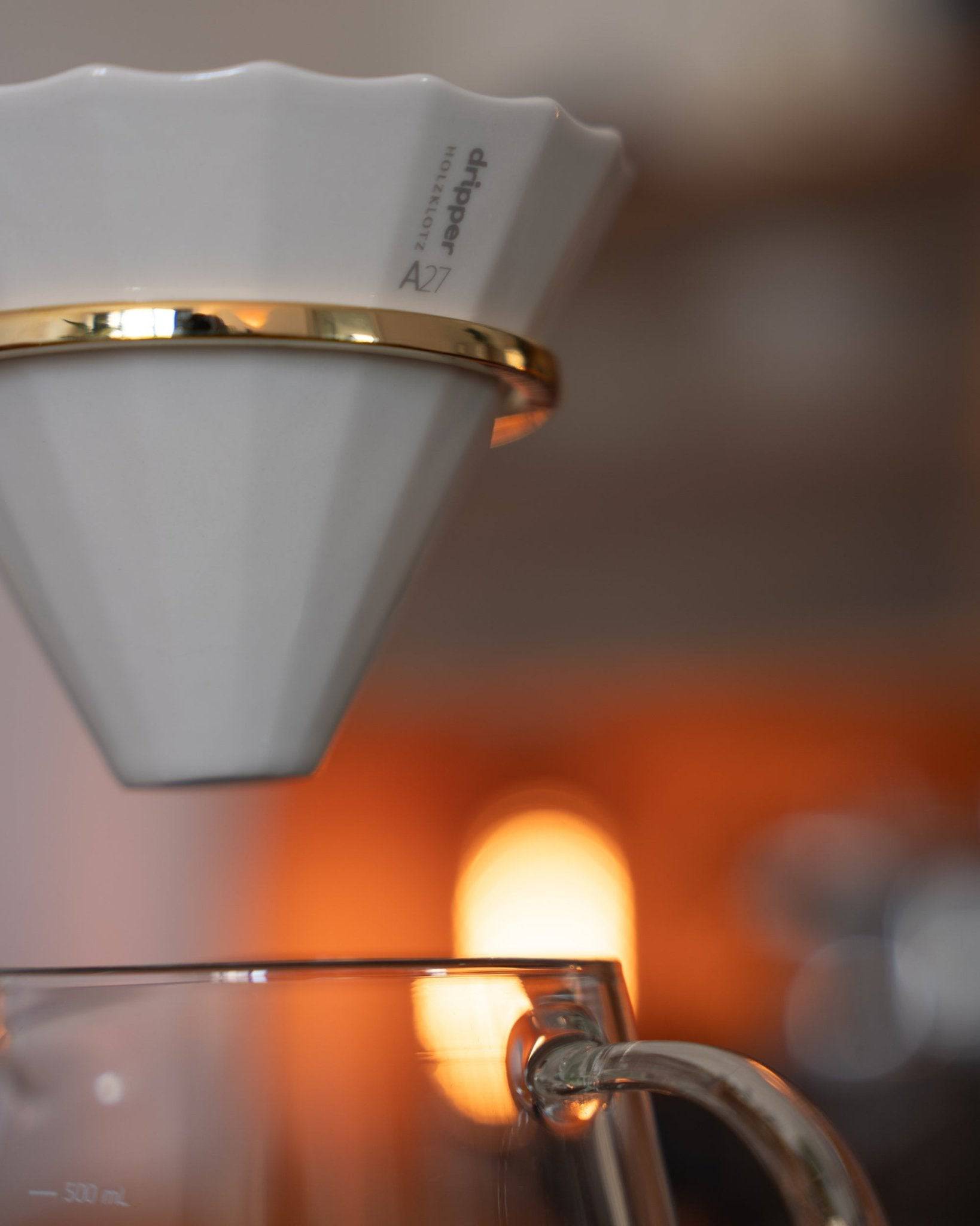 White ceramic pour-over coffee dripper with gold rim on glass carafe, showcasing Holzklotz&