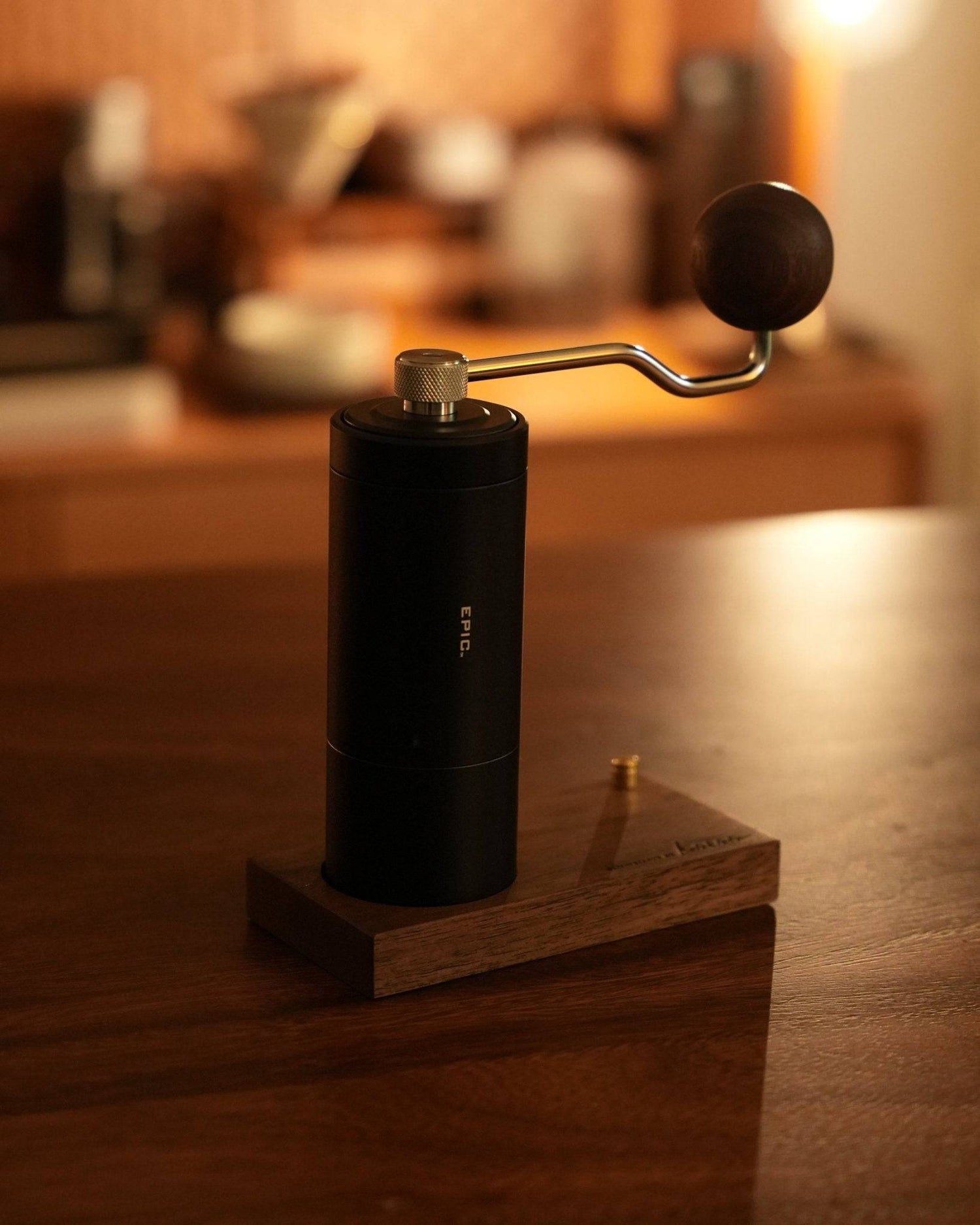 Stylish black hand coffee grinder with metal handle on wooden stand, warm lighting, kitchen countertop setting, coffee equipment in background