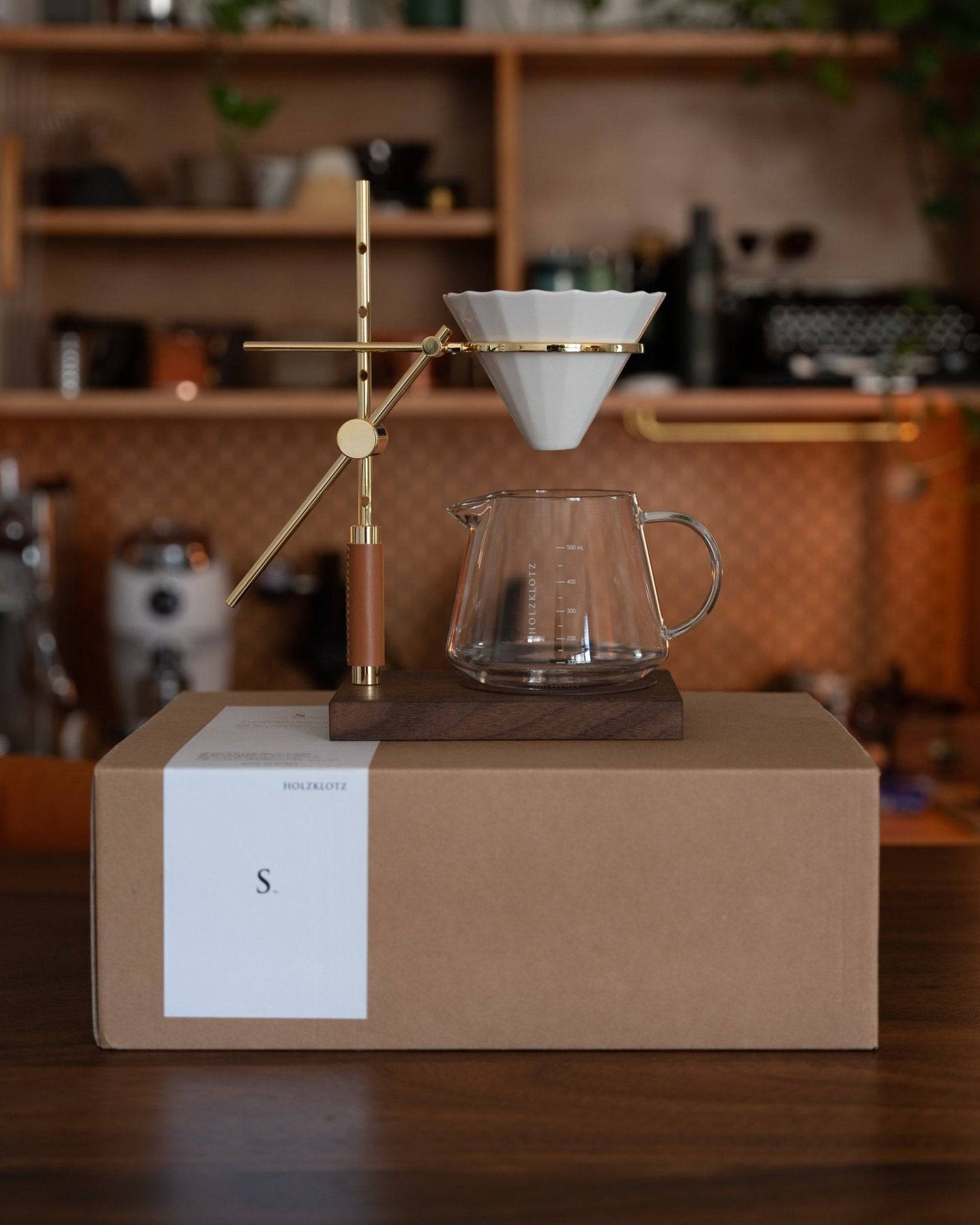 Holzklotz pour-over coffee set featuring gold-plated stand, ceramic filter, and glass carafe on wooden base, displayed on cardboard box in cozy kitchen setting