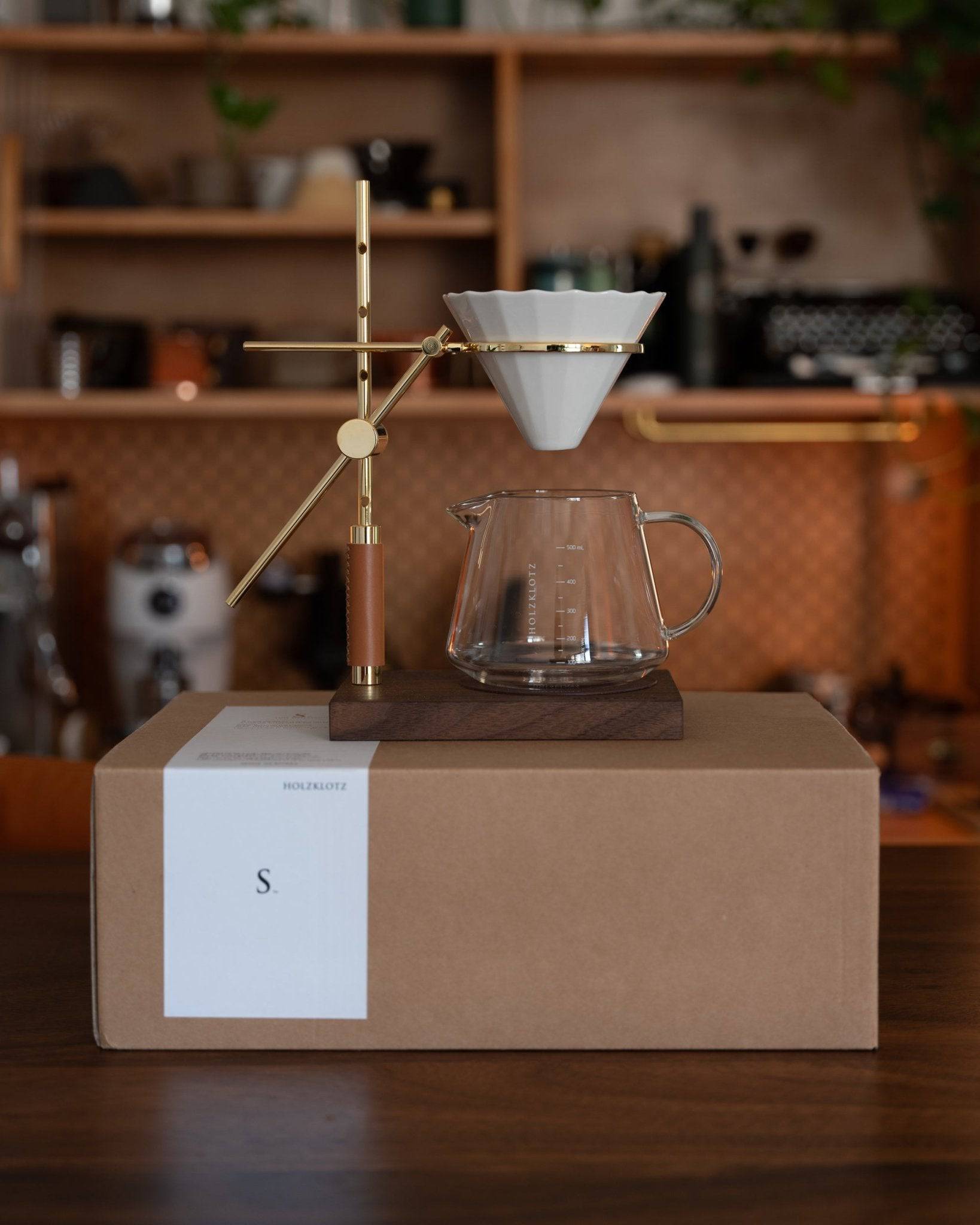 Holzklotz pour-over coffee set featuring gold-plated stand, ceramic filter, and glass carafe on wooden base, displayed on cardboard box in cozy kitchen setting