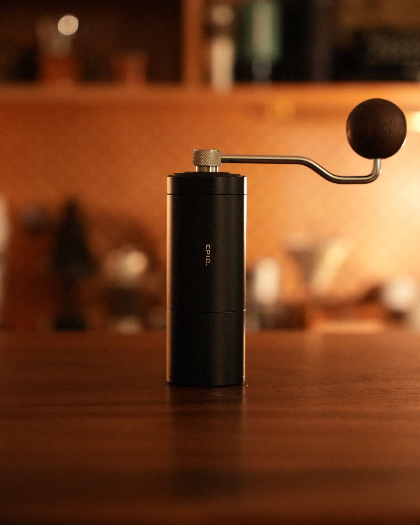 Black Holzklotz manual coffee grinder with sleek cylindrical design and crank handle, placed on wooden surface in warm, ambient lighting. Blurred background suggests kitchen or cafe setting.