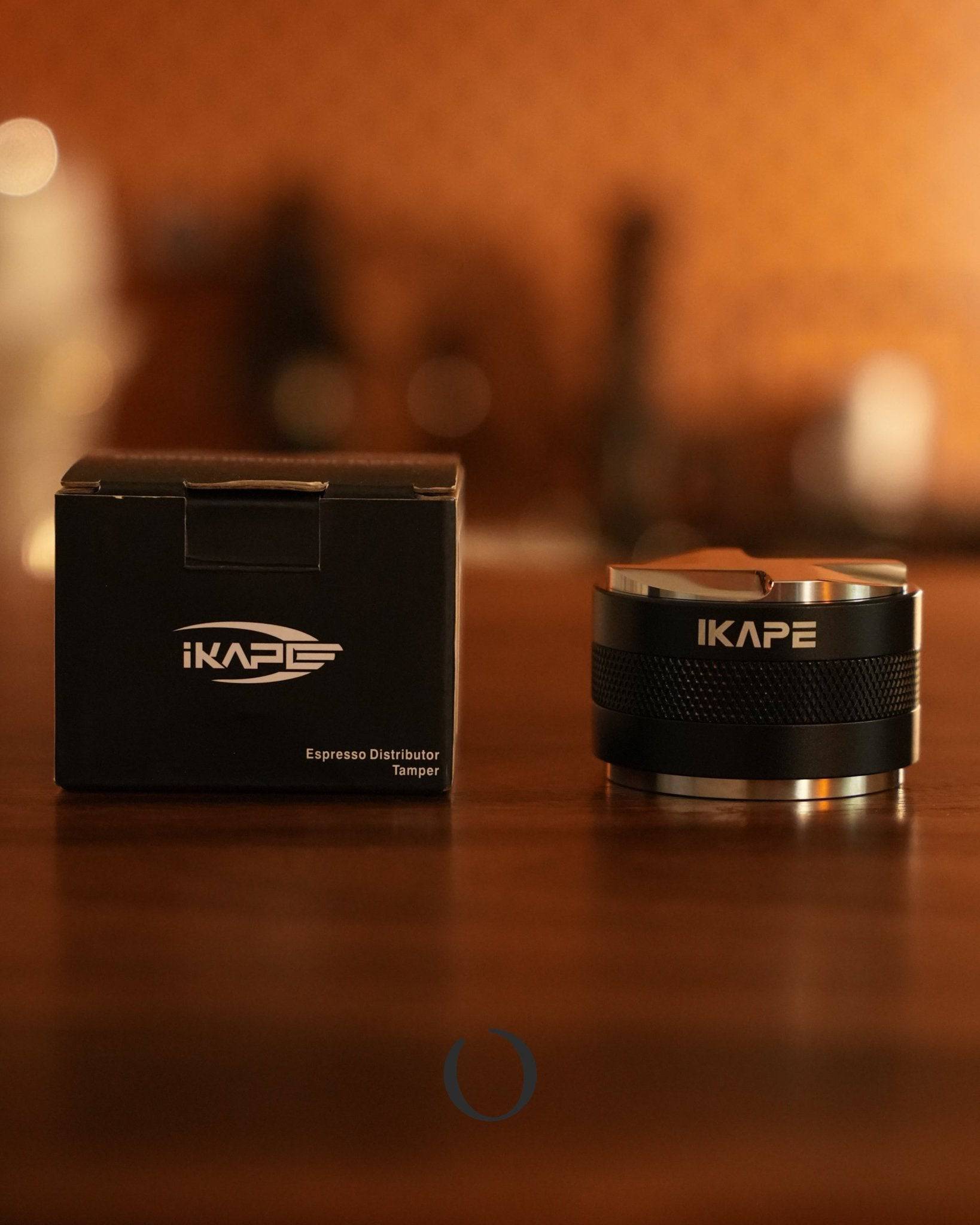 IKAPE Espresso Distributor and Tamper 2-in-1 tool with black packaging box on wooden surface, warm ambient lighting, coffee equipment silhouettes in background, professional barista accessory for consistent coffee grounds distribution and tamping