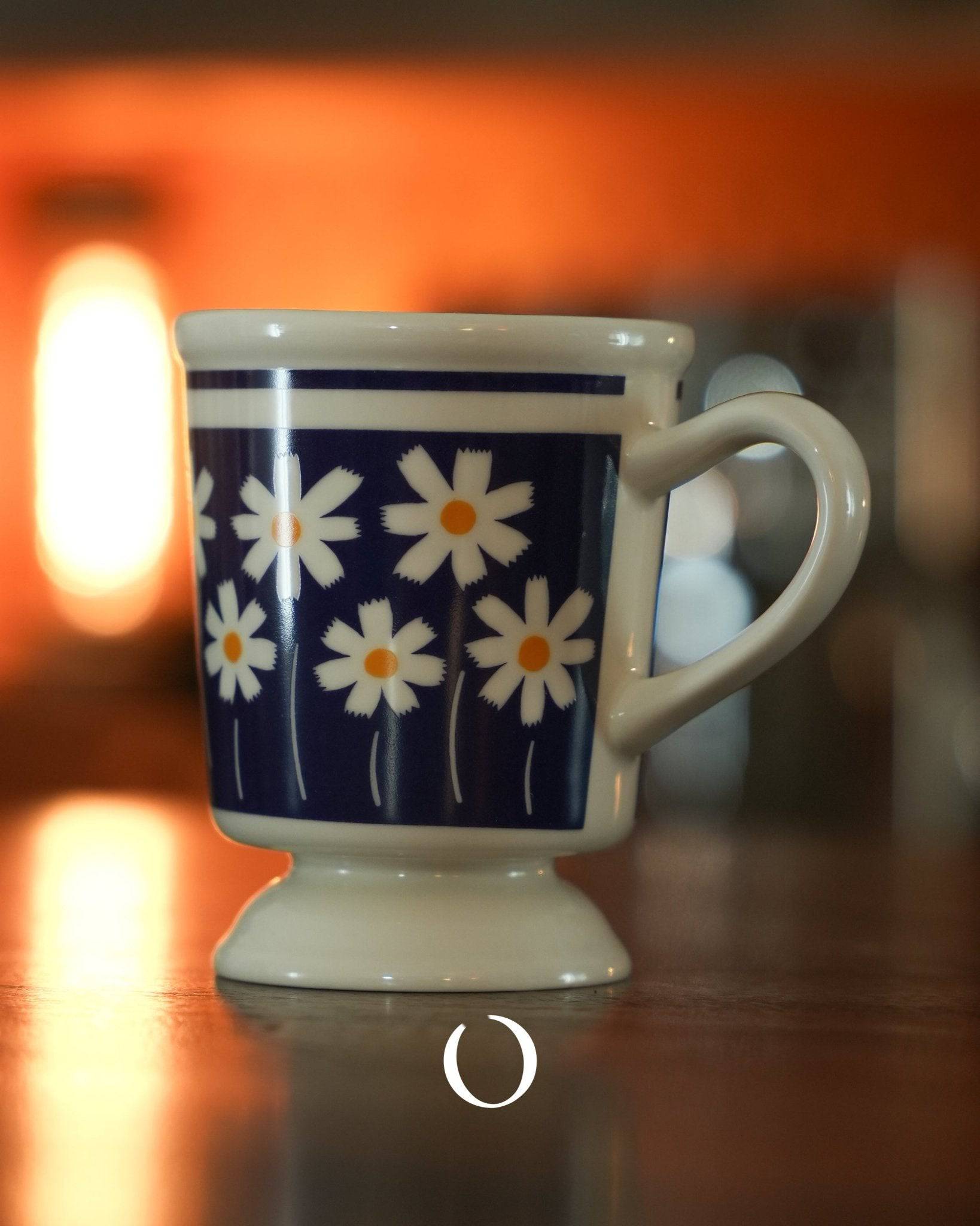 Vintage-style ceramic coffee cup with white daisy pattern on deep blue background, showcasing Showa era design, placed on reflective surface with warm orange glow in background