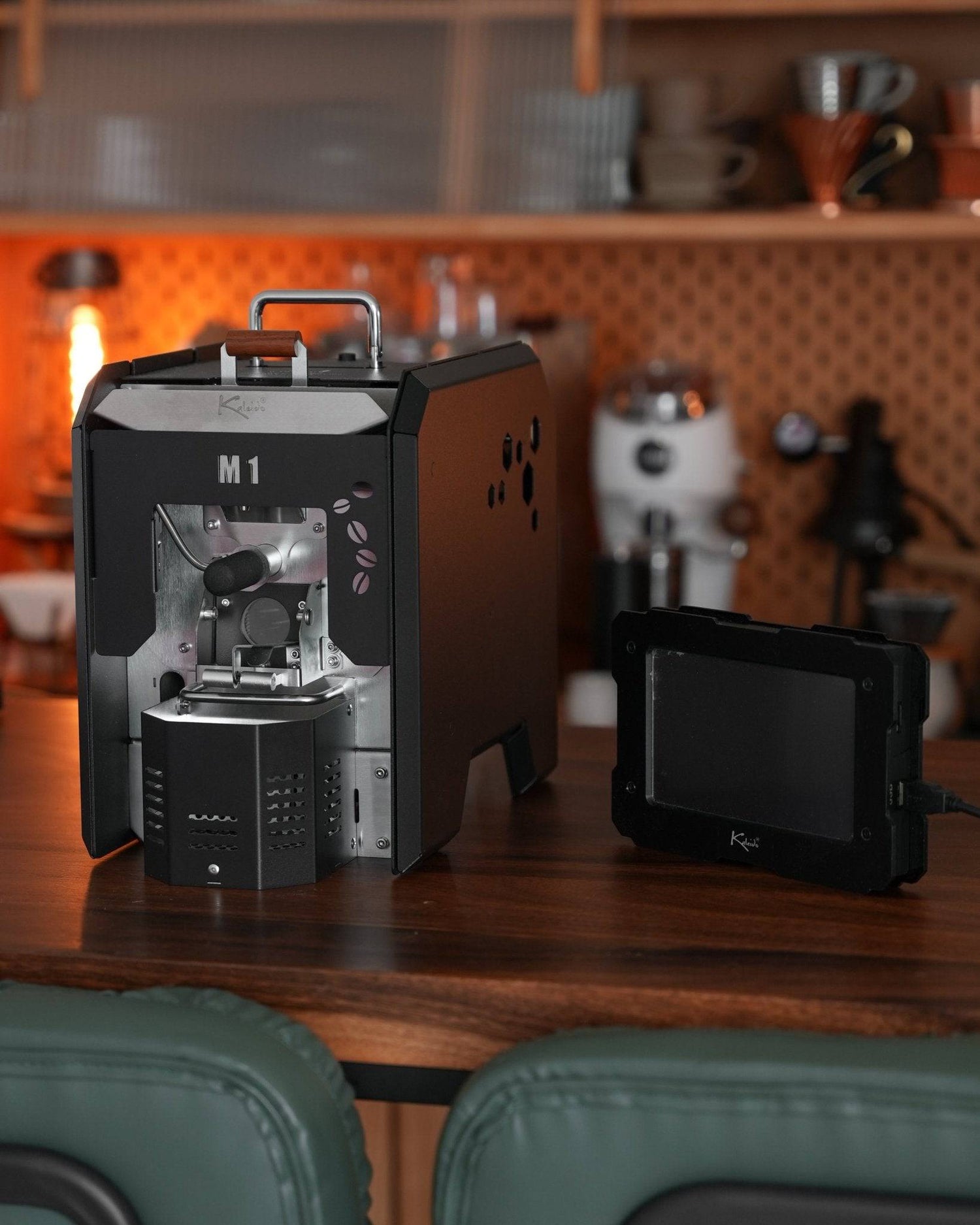 KALEIDO M1 coffee roaster on wooden table in kitchen, with touchscreen display, visible roasting chamber, and cozy orange background