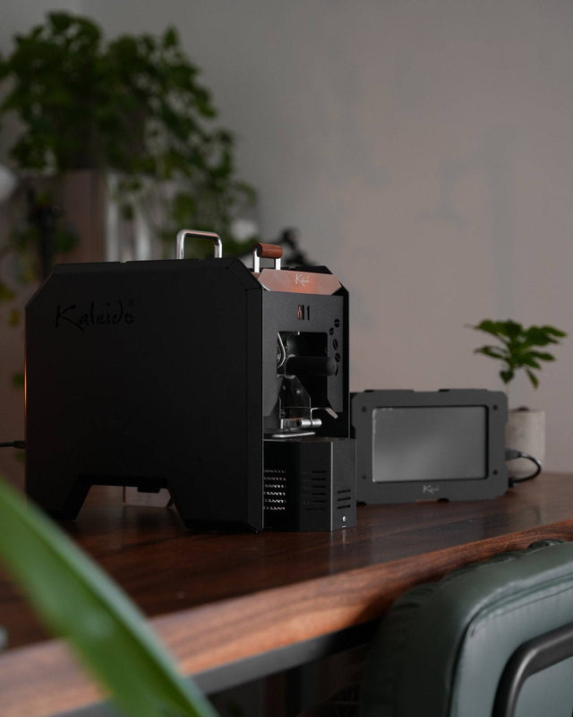 KALEIDO M1 coffee roaster on wooden table, sleek black design with digital display, surrounded by plants in home setting