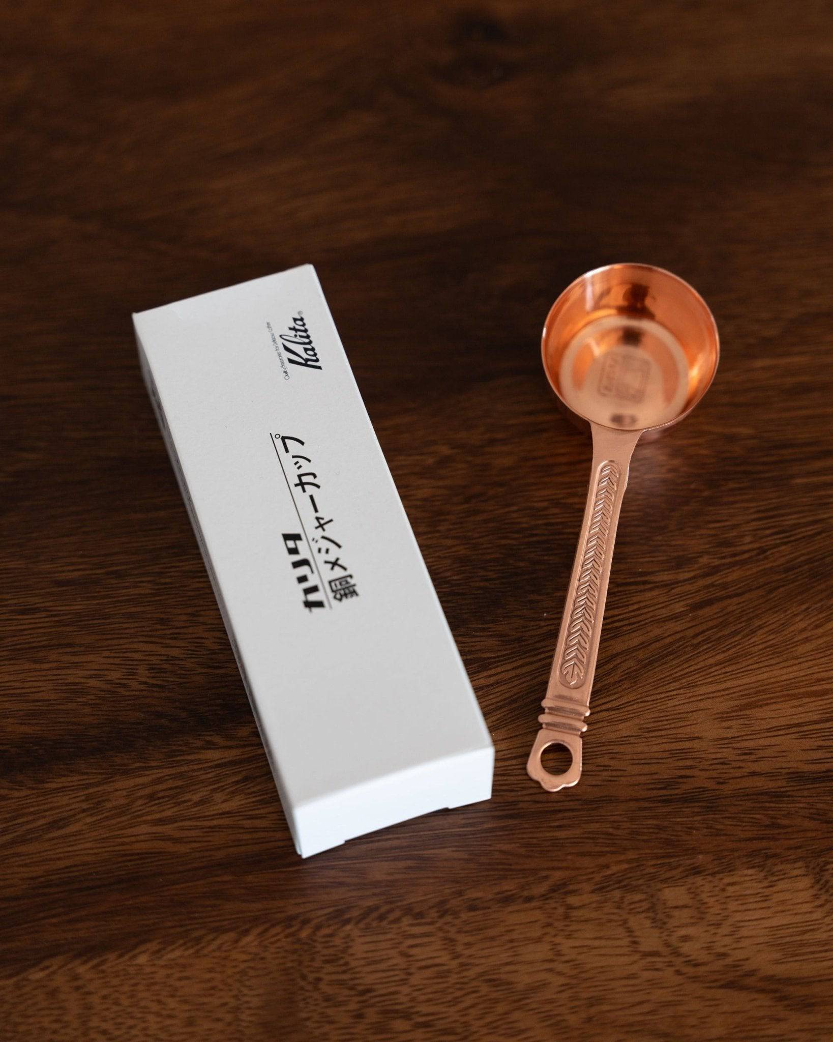 Kalita copper coffee scoop with laurel pattern, 10g capacity, next to its white branded packaging on wood surface