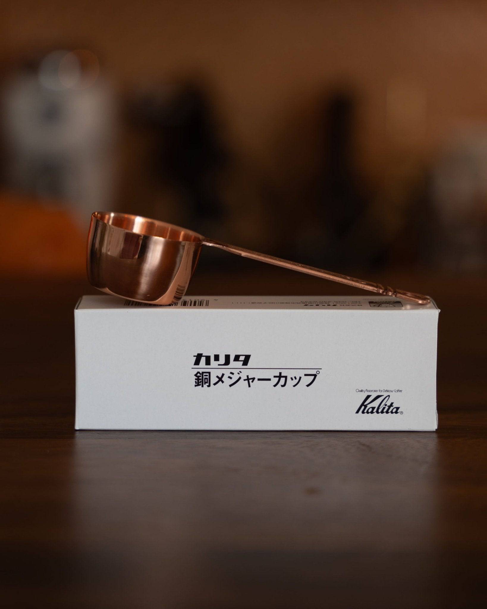 Kalita copper coffee scoop with laurel wreath pattern, 10g capacity, resting on its product box, showcasing elegant design and premium quality for precise coffee measurement