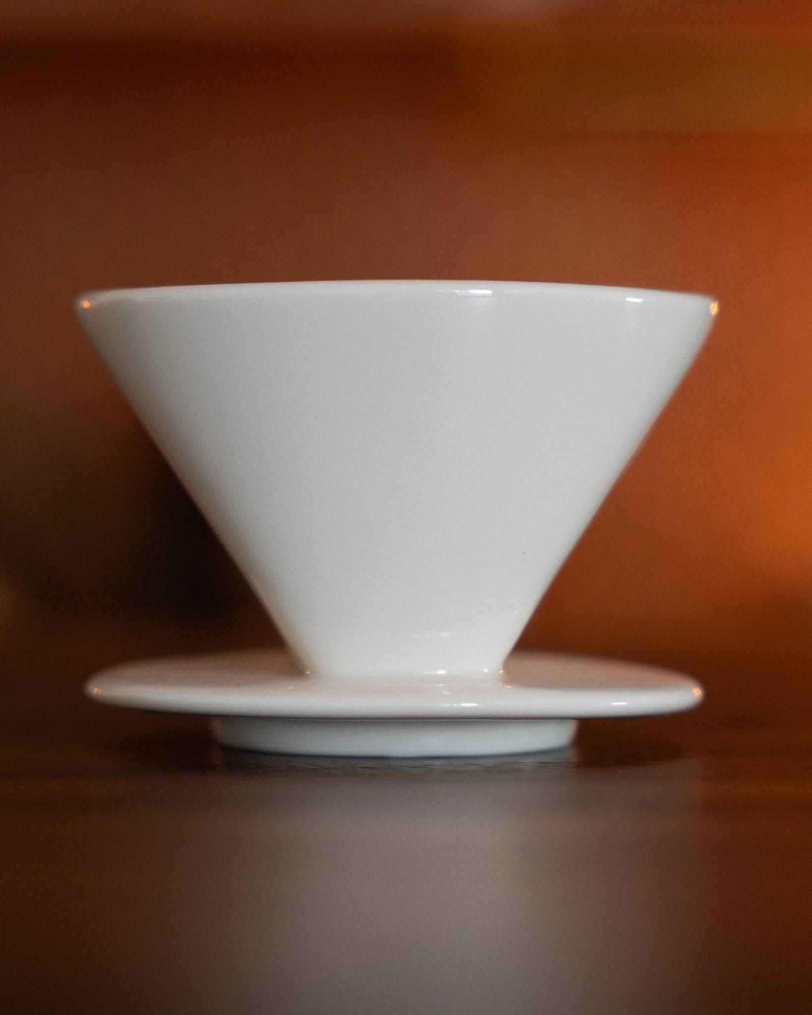 White Kalita ceramic pour-over coffee dripper, cone-shaped with a flat base, showcasing its elegant design and high-quality craftsmanship against a warm brown background