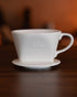 Kalita ceramic pour-over coffee dripper 101 in white, featuring a conical shape and handle, placed on a wooden surface with warm lighting, showcasing its elegant design for brewing specialty coffee