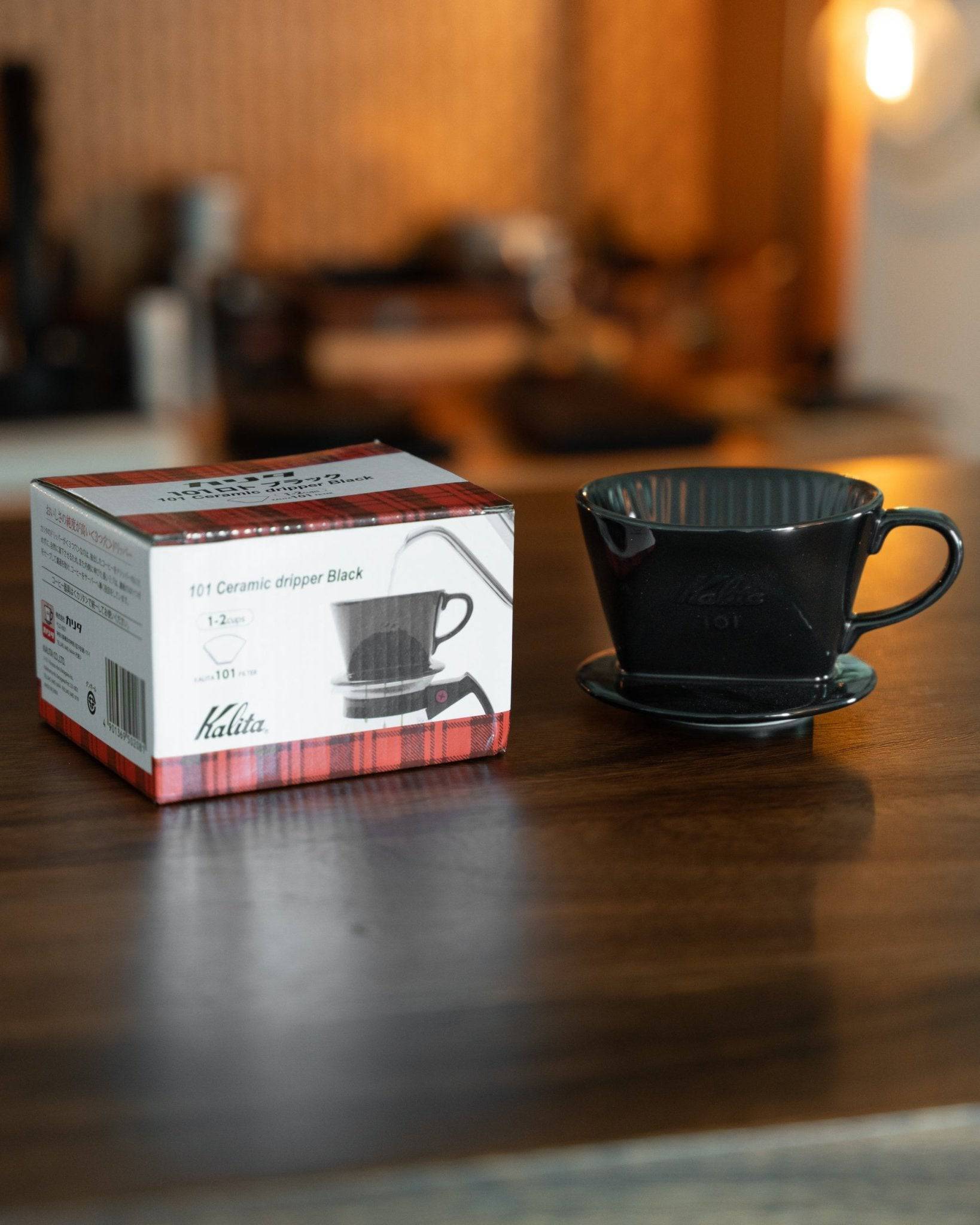 Kalita 101 black ceramic coffee dripper with product box on wooden table, coffee brewing equipment in background, warm lighting