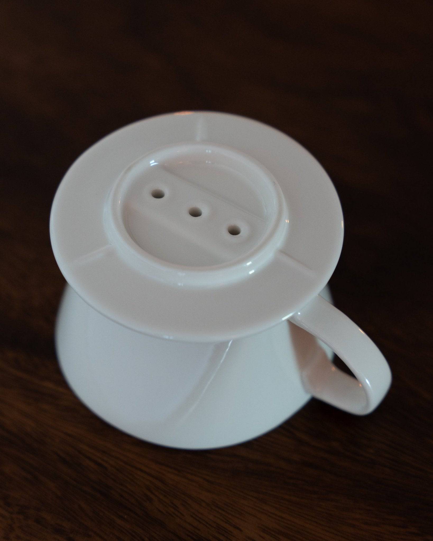 White Kalita ceramic coffee dripper with three holes, conical shape, and handle on dark wooden surface. Japanese-made pour-over coffee filter for 1-2 cups, showcasing unique design for optimal brewing.