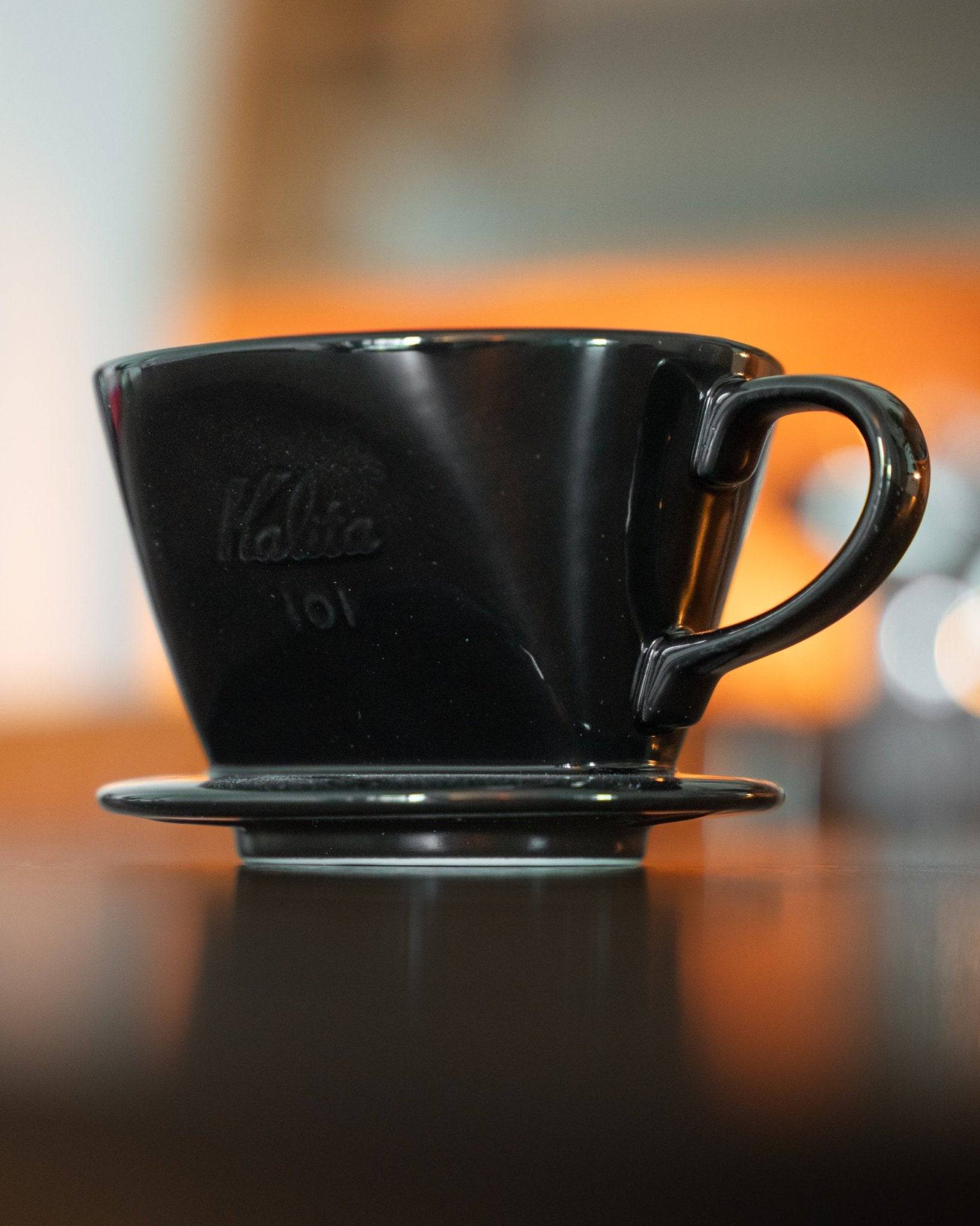 Black Kalita 101 ceramic coffee dripper on reflective surface with orange background, showcasing its sleek design and unique shape for optimal coffee brewing