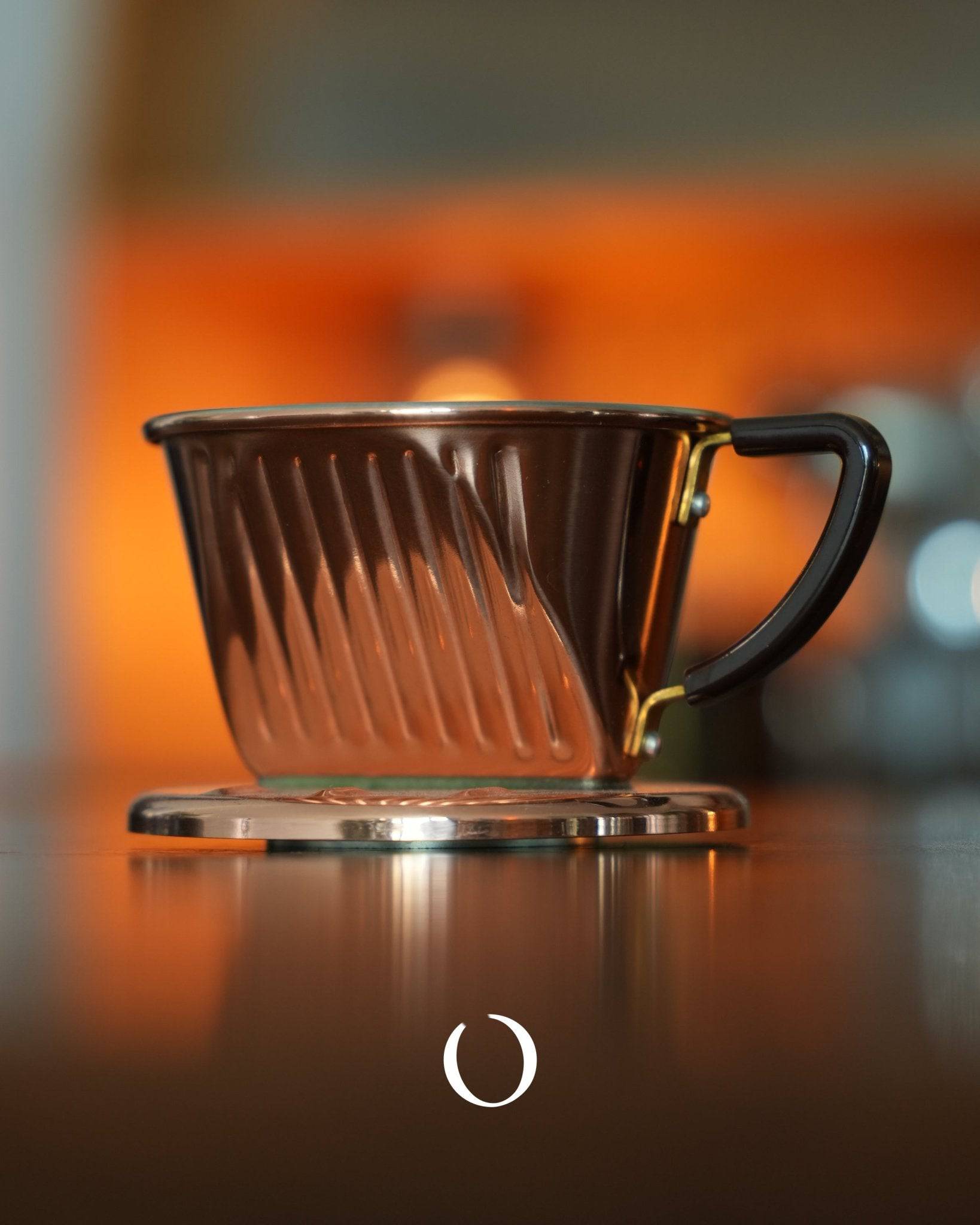 KALITA 101-CU copper coffee dripper with three-hole design on orange background, showcasing its unique spiral ridges and handle, sitting on a reflective surface for optimal brewing and aesthetic appeal