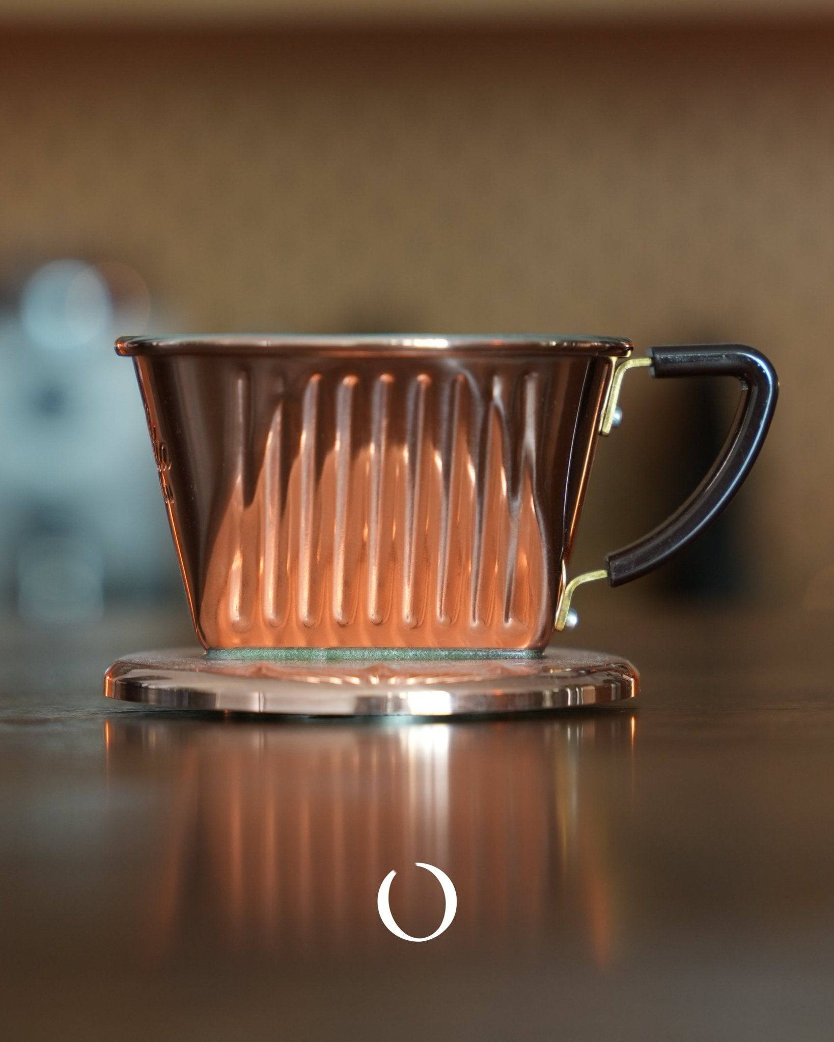 KALITA copper pour-over coffee dripper with ribbed design and black handle, sitting on reflective surface, showcasing elegant craftsmanship and premium quality for coffee brewing