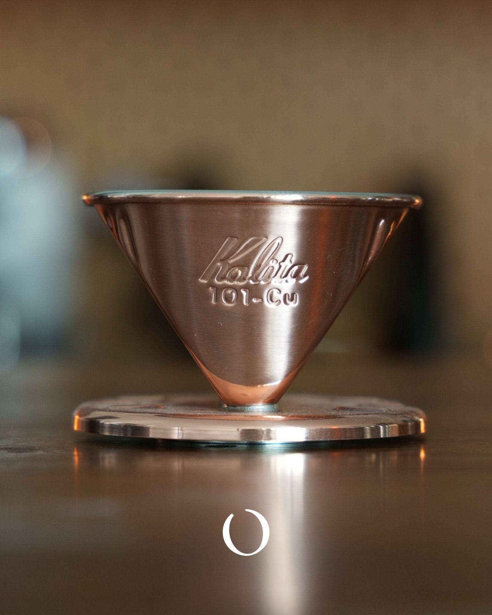 Kalita 101-CU copper coffee dripper with three-hole design, showcasing its conical shape and shiny copper finish on a wooden surface, highlighting its artisanal craftsmanship and heat-conducting properties for optimal coffee brewing.